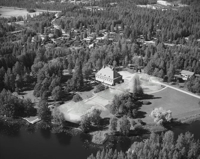 Aerial view of a park with a large building, tennis courts, trees, and a body of water, surrounded by a forest and residential area.