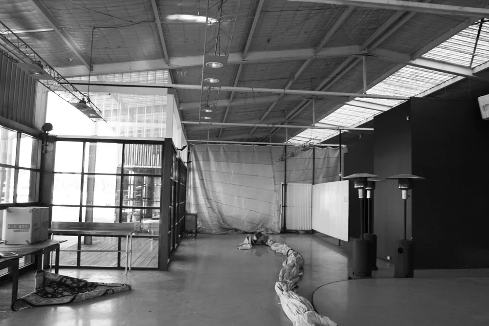 Interior of a building with large windows, a table with boxes, a heater, and some fabric or tarp on the floor, suggesting it might be under construction or renovation.