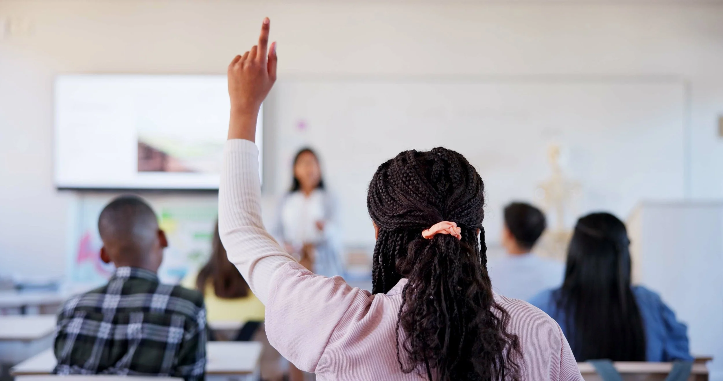 A classroom scene with students, one of whom is raising her hand to ask or answer a question, while the teacher stands at the front near a whiteboard or screen.