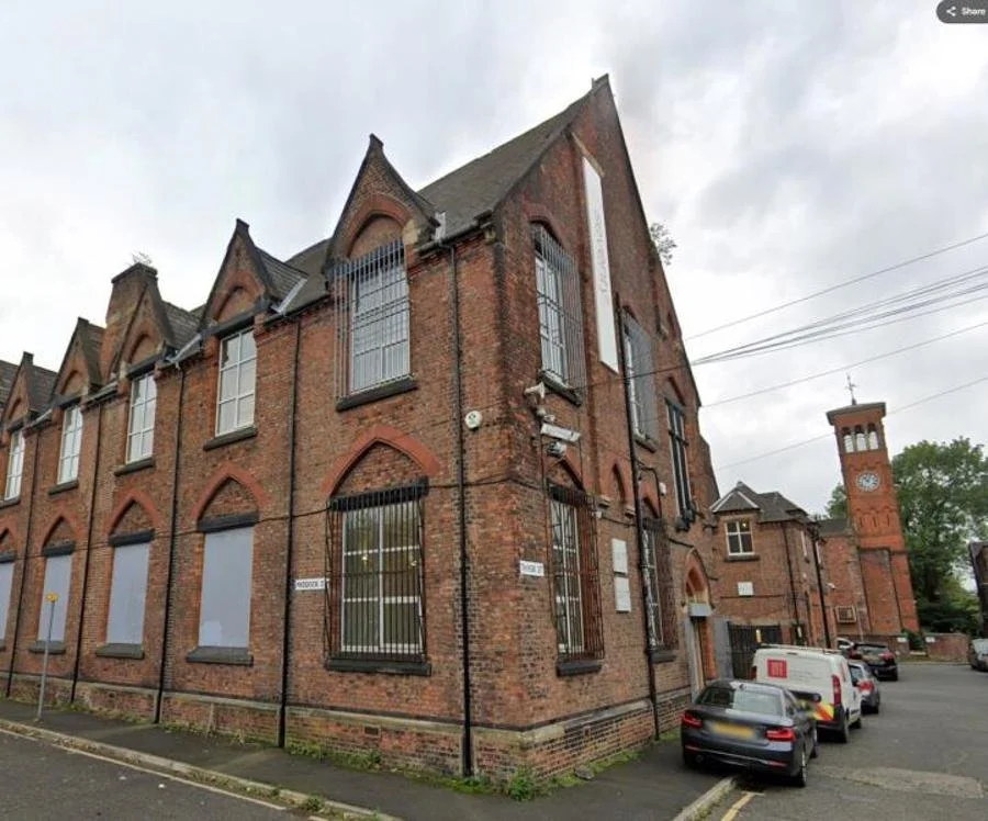 Brick building with barred windows and a clock tower in the background, parked cars along the street, overcast sky.