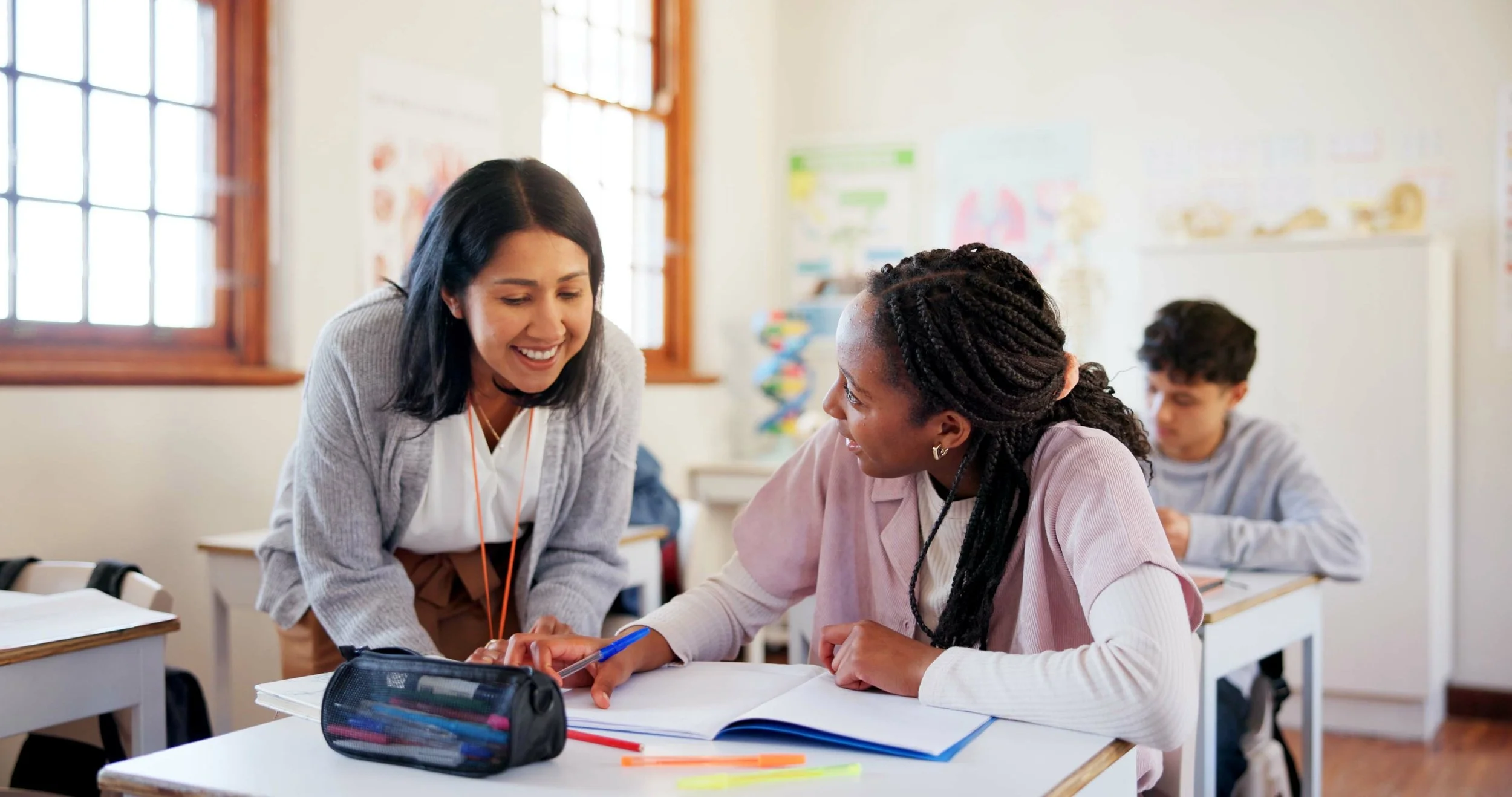 Teacher assisting a student with her homework in a classroom with other students visible in the background.