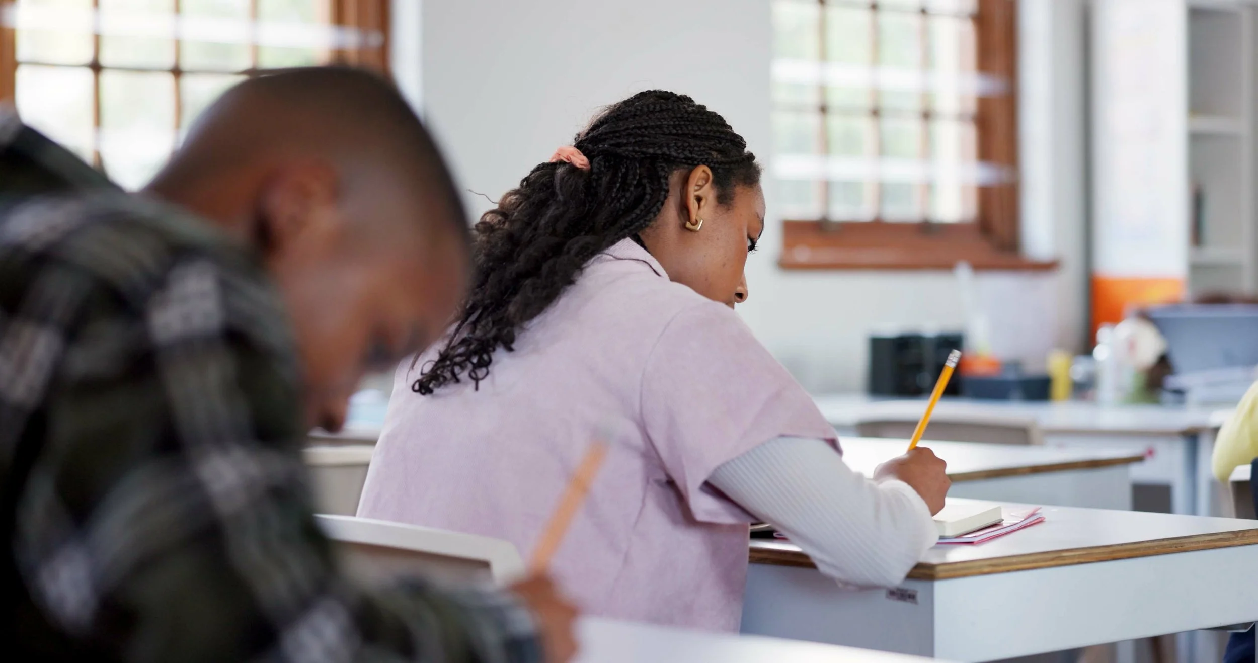 Students sitting at desks in a classroom, writing notes, with a woman in a pink top taking notes and students in the background near windows.