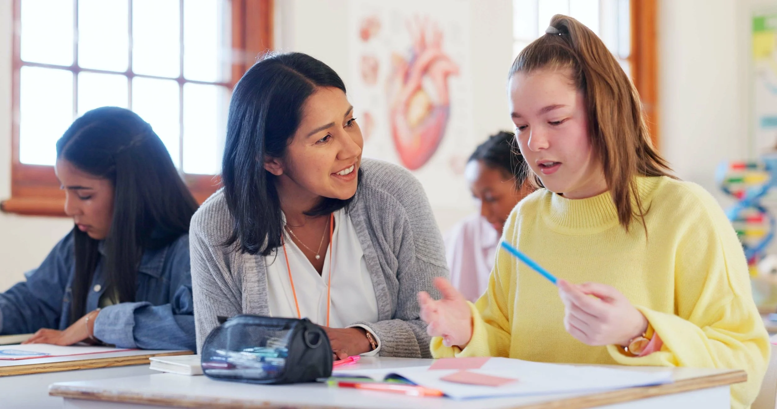 Teacher assisting female student working on a project in classroom with educational posters and windows in background.