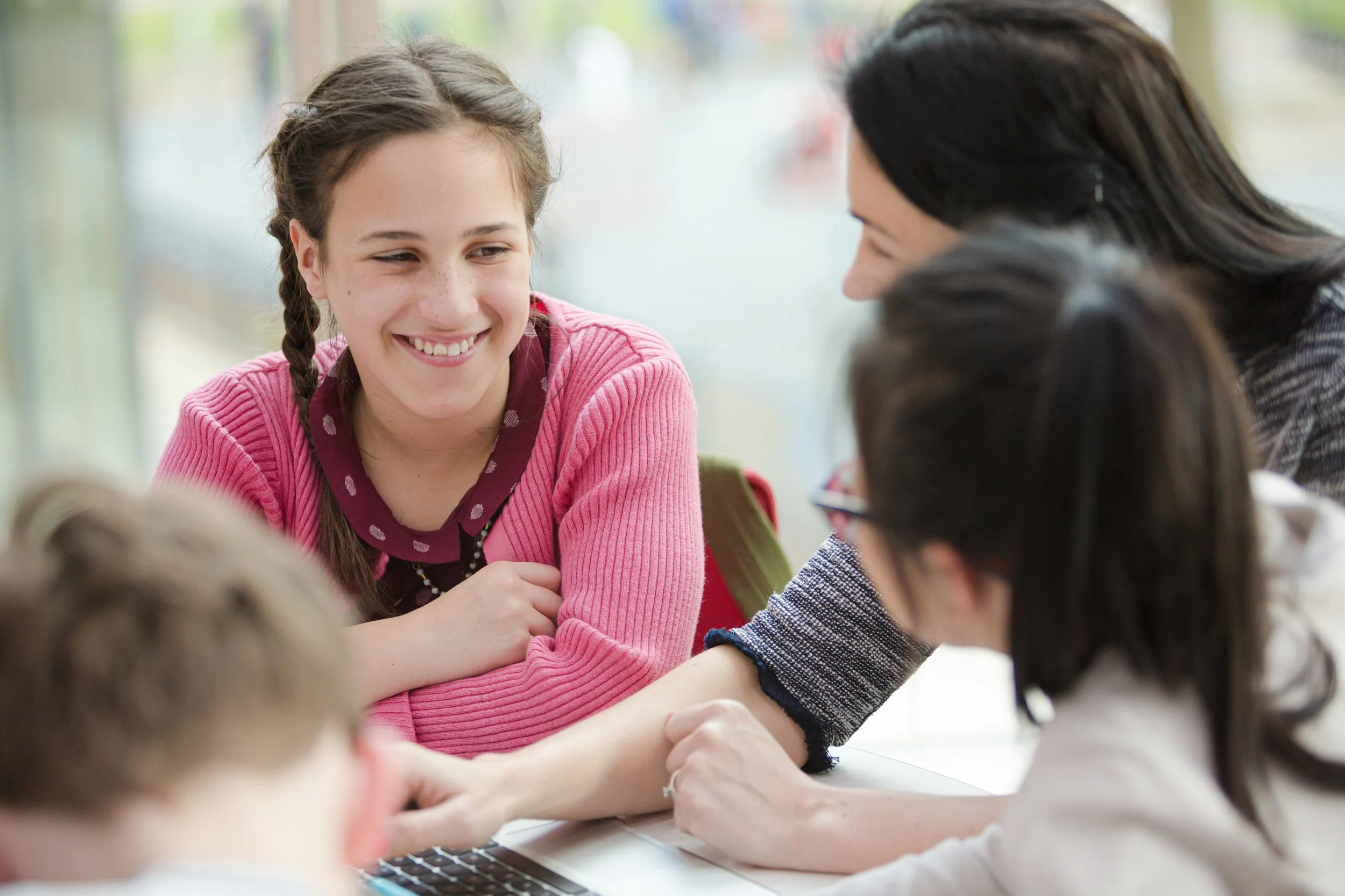 A young girl with braided hair smiling, sitting at a table with her arms crossed, engaging in conversation with other children and a woman in a classroom or similar setting.