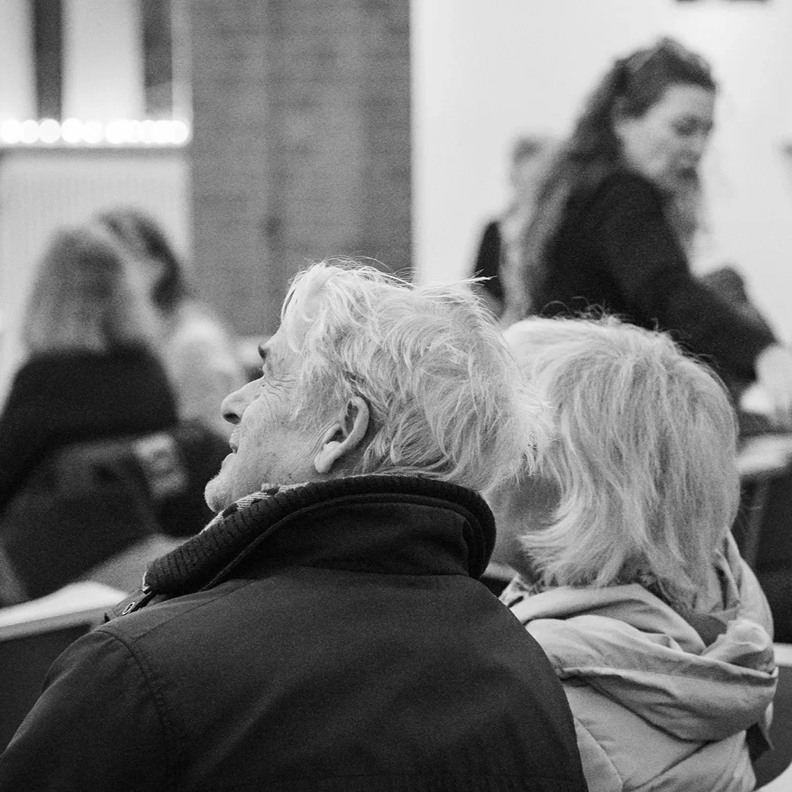 A black and white photo of two people sitting in a relaxed social setting, just before a concert, observing their surroundings. Other people gathering in the background.
