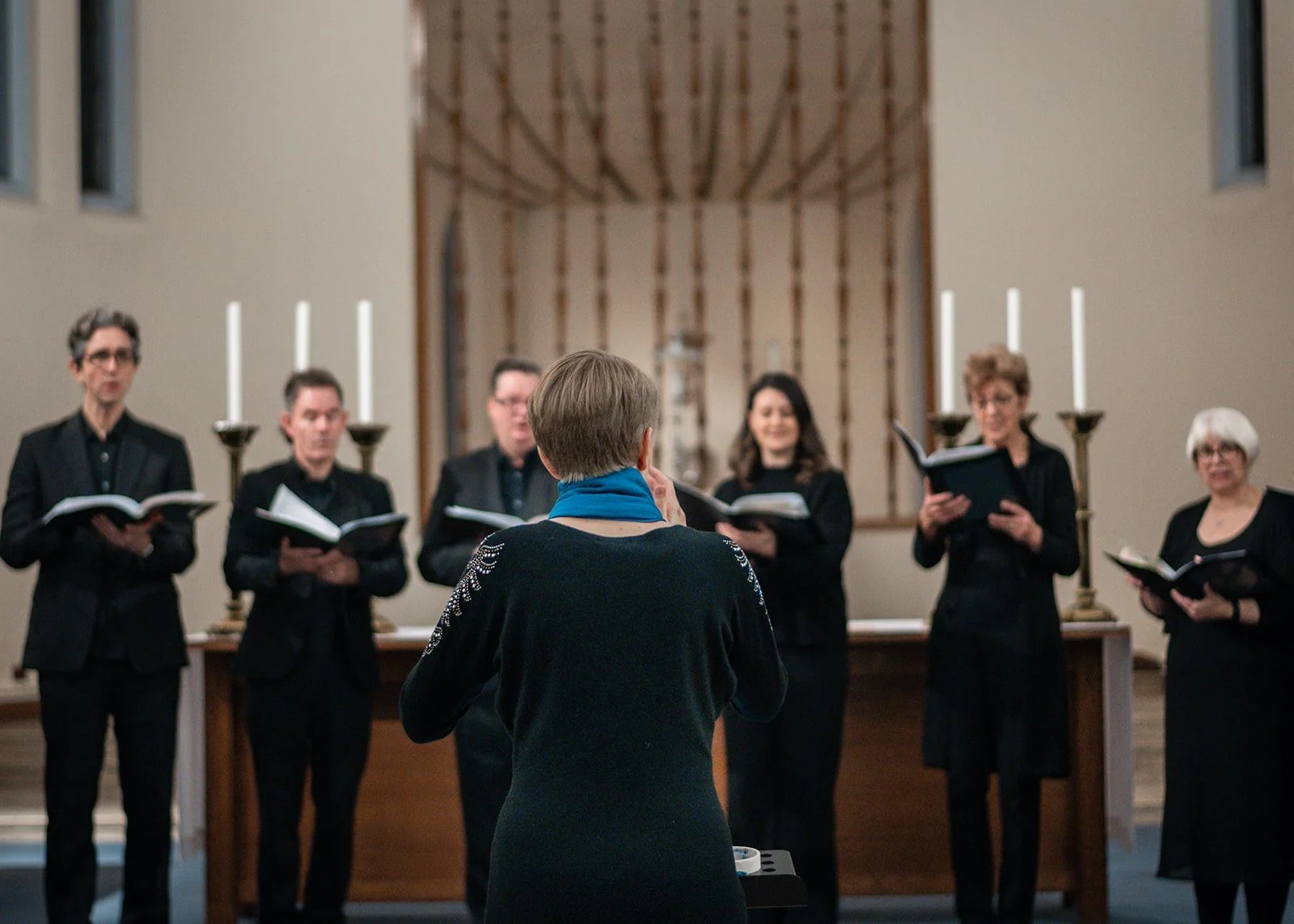 A choir practicing in front of a conductor in a church, with lit candles and a wooden screen in the background