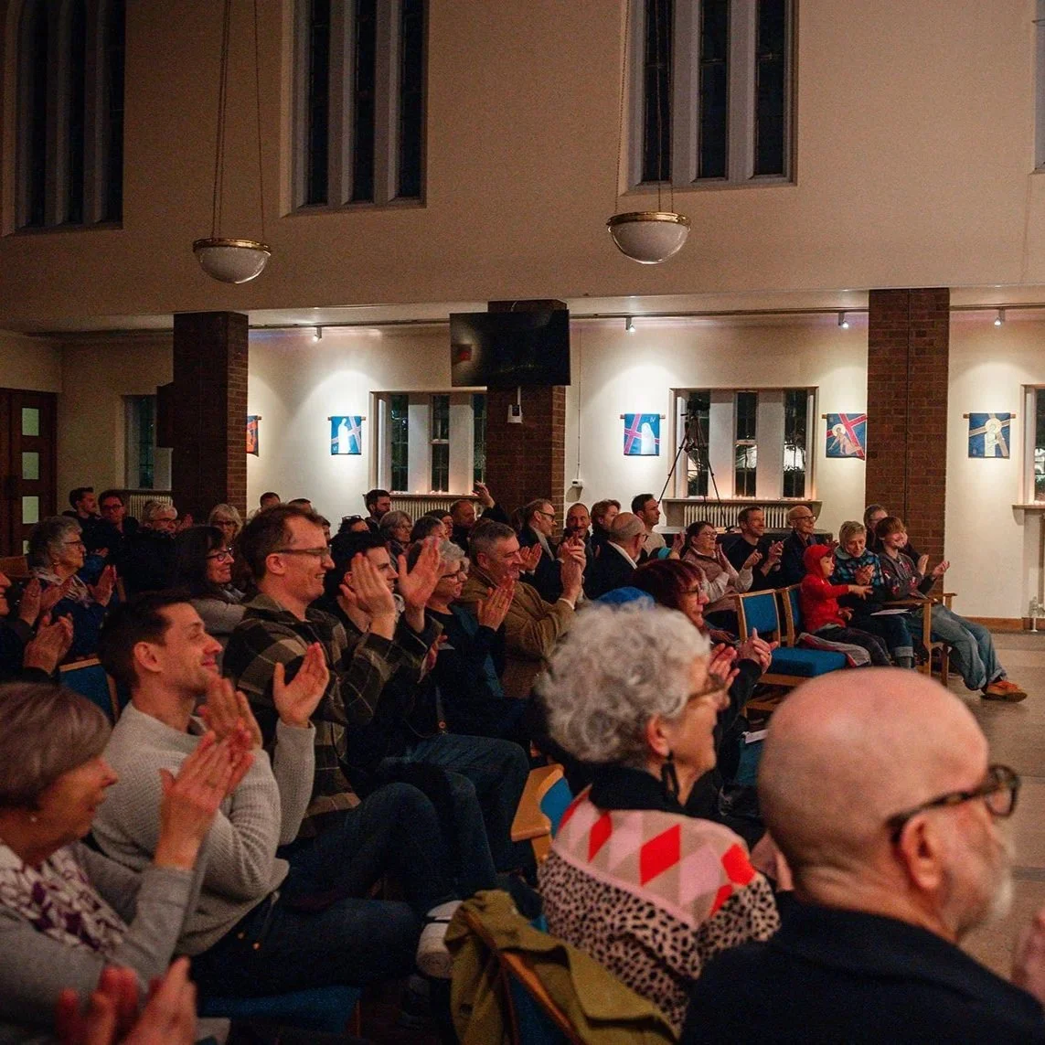 A seated audience of all ages, listening to a performance in a church venue with artwork on the walls. Some people are smiling.
