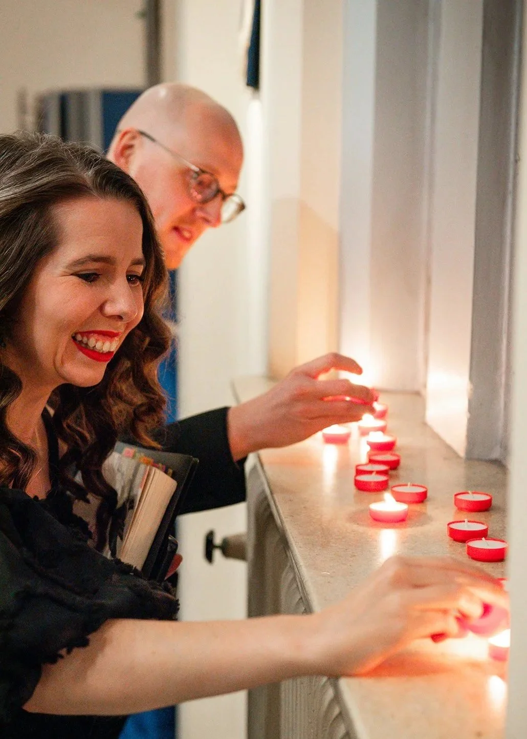 Two people dressed in dark clothing are lighting red tea-lights on a church windowsill. The woman has long dark dark hair and is wearing red lipstick, and a man with a bald head wearing glasses. They look excited.