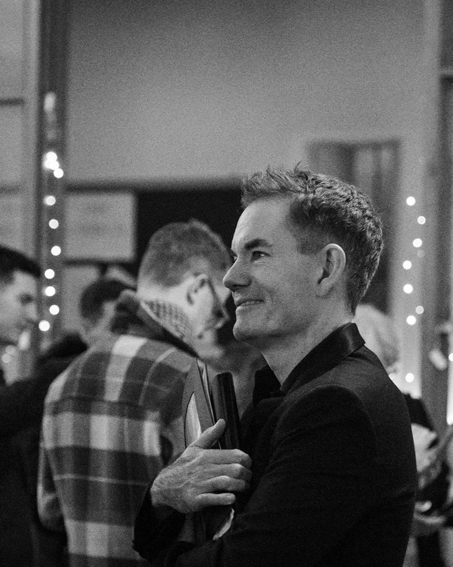 A man smiling, dressed in a black suit jacket, holding a folder of choir music, at a post-concert social gathering, with other people in the background
