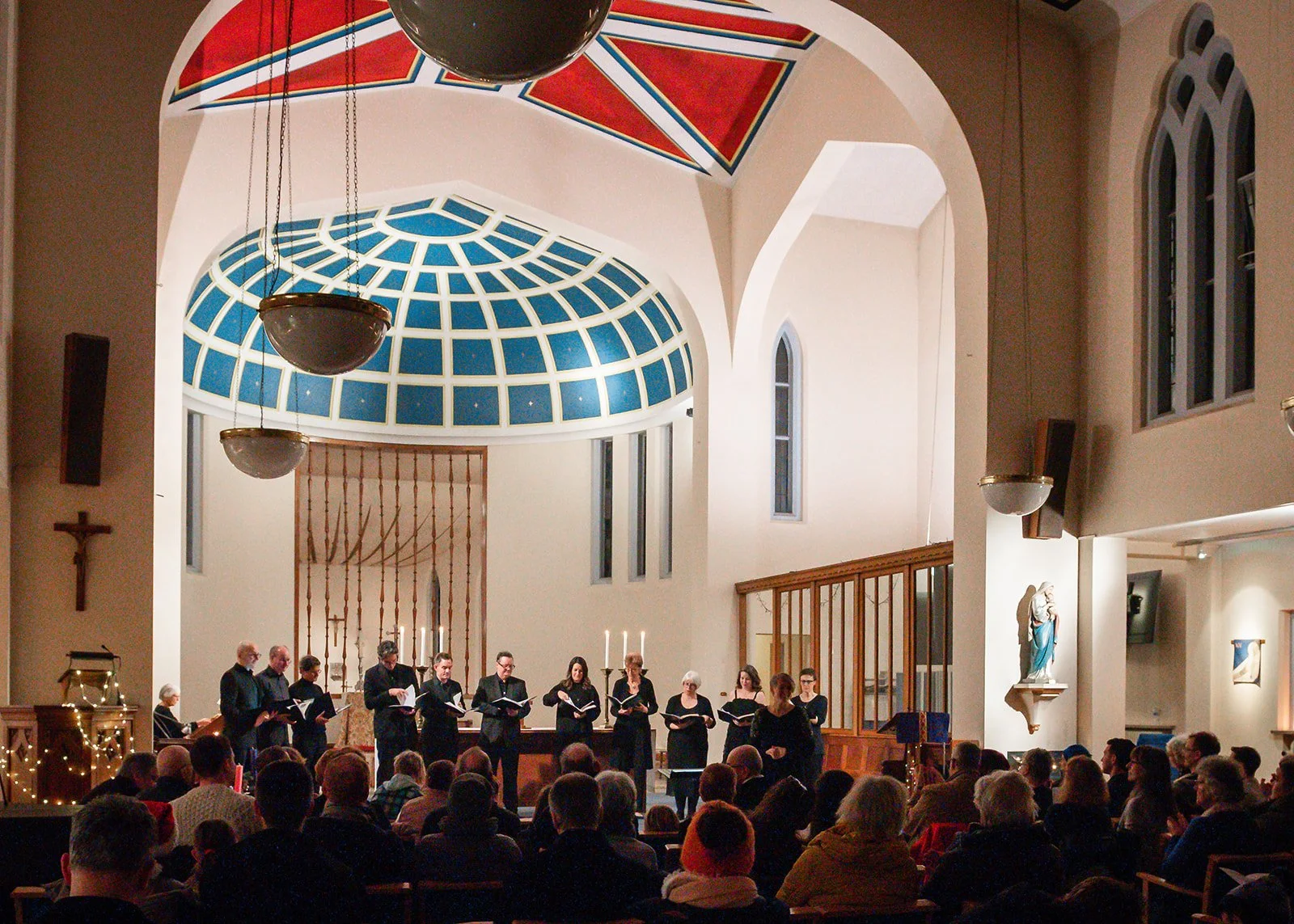 Choir performing in front of the altar of a church, beneath a well-lit blue and red domed-ceiling, with an audience seated in chairs amongst candlelight.