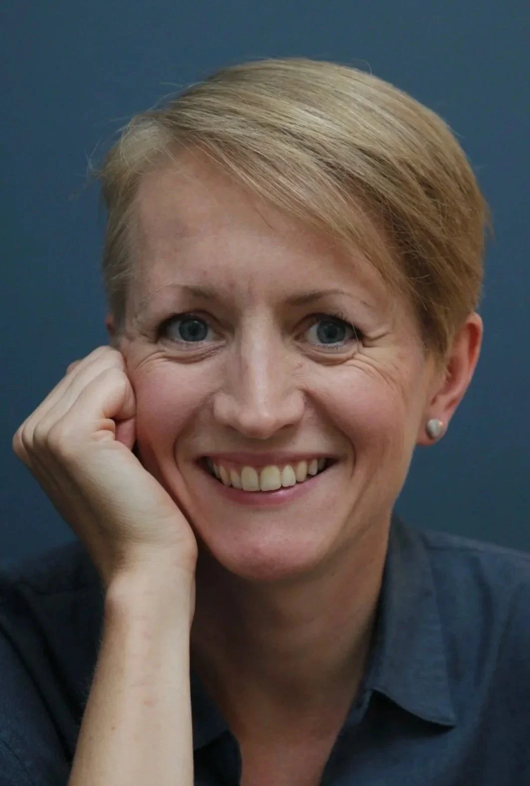 Close-up of smiling woman with short blonde hair, blue eyes, wearing a dark shirt, resting her face on her hand against a dark background.