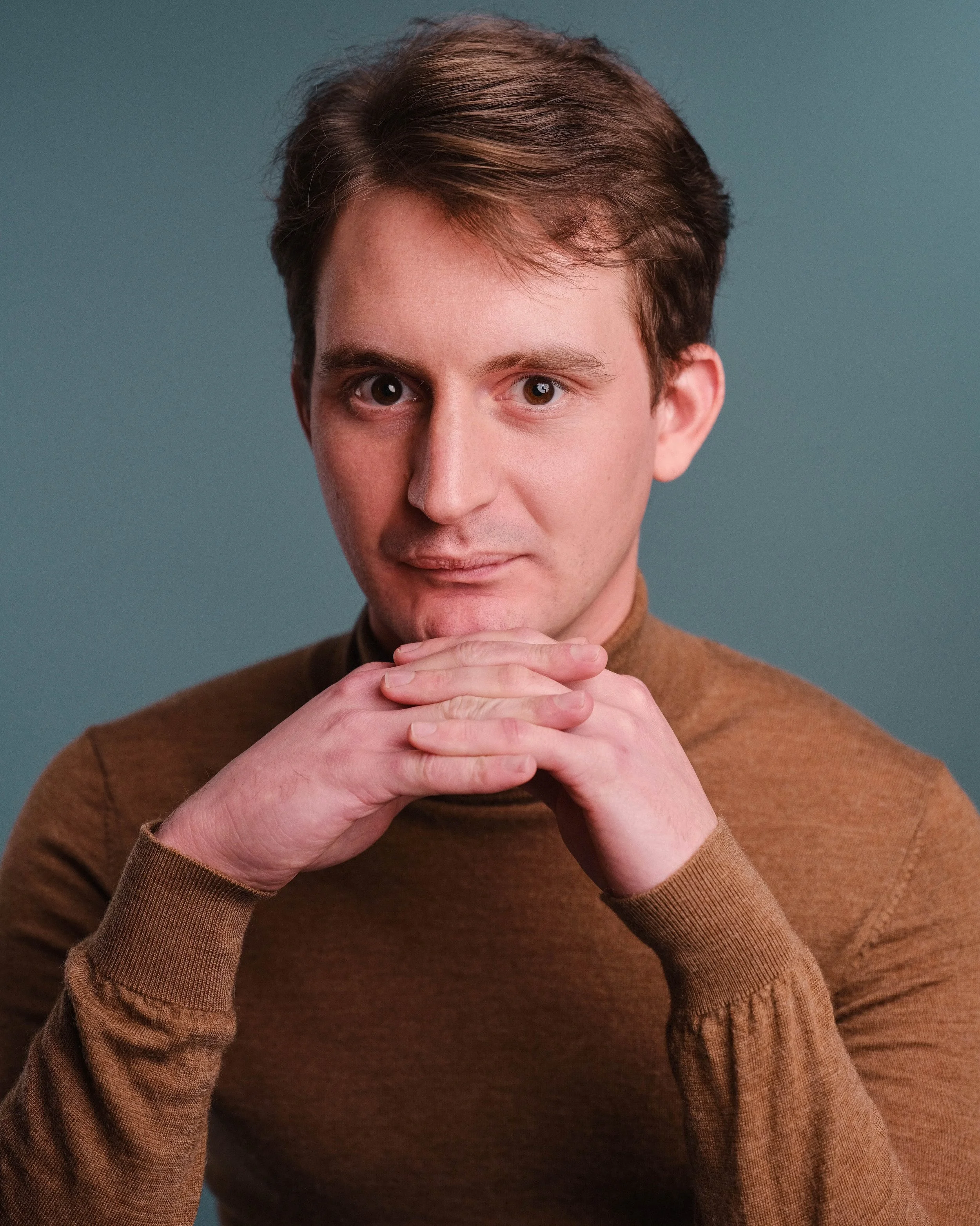 A young man with brown hair and brown eyes, wearing a brown sweater, looking directly at the camera with hands clasped under his chin against a blue background.