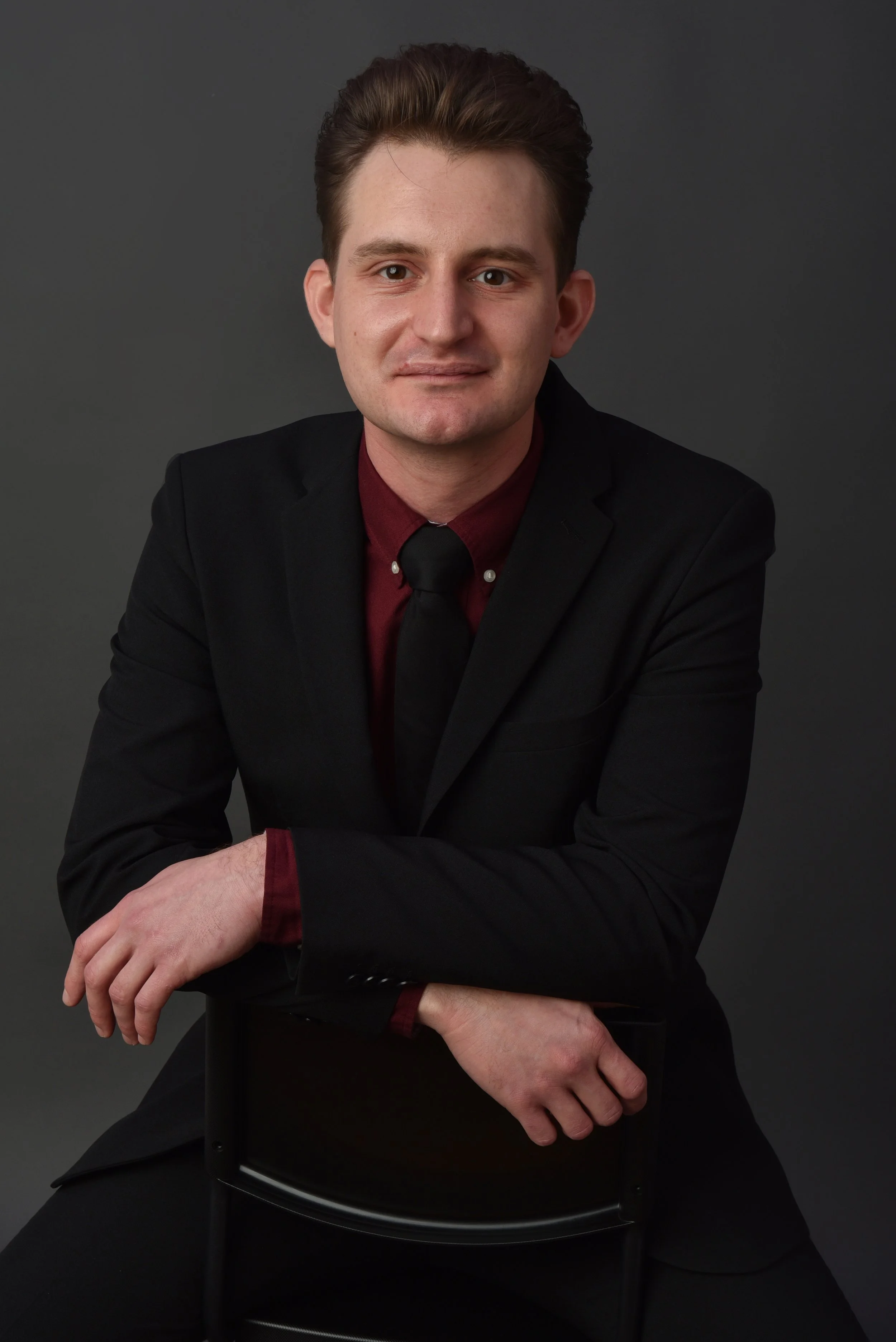 A young man in a black suit, maroon shirt, and black tie sitting against a dark background.