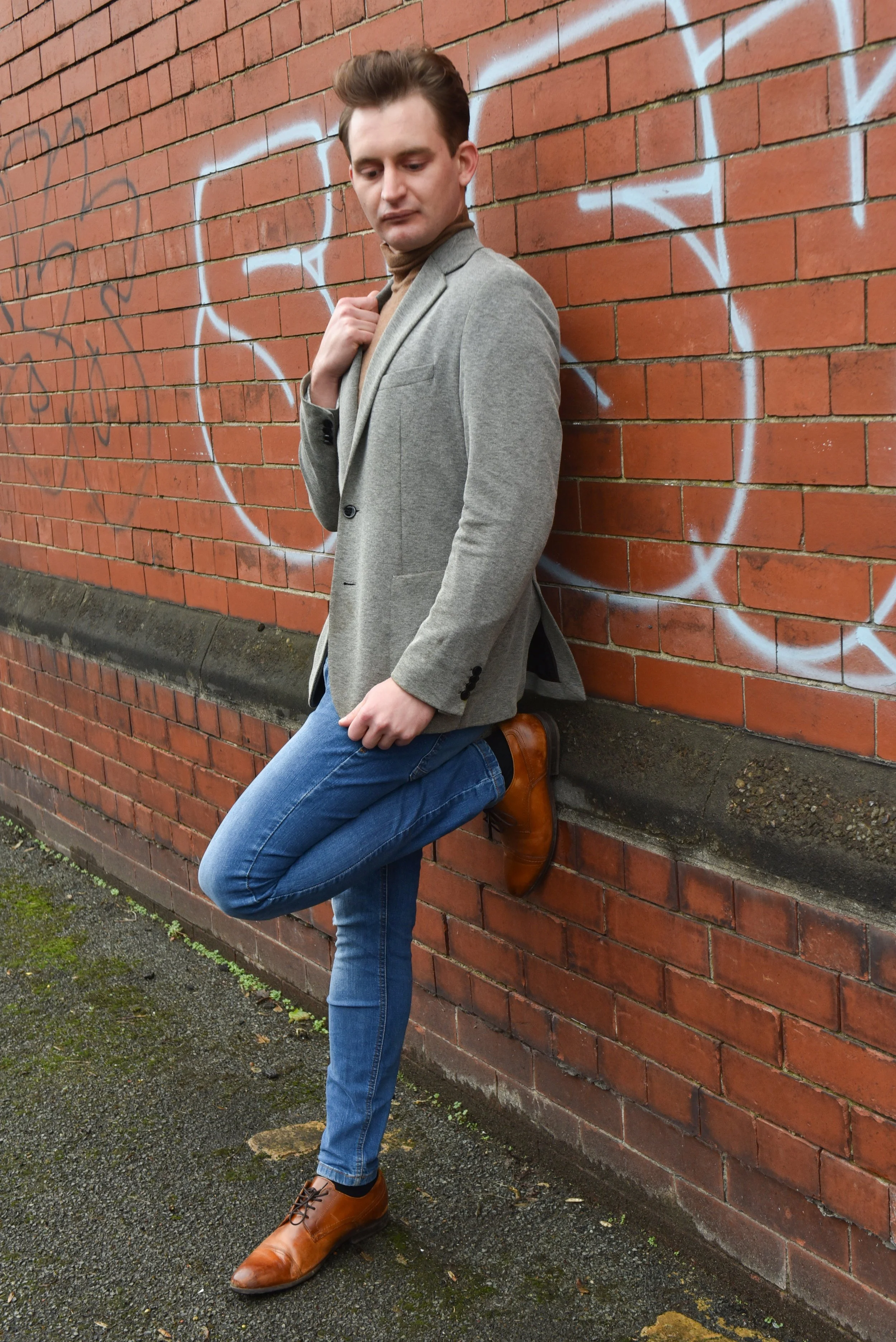 A young man leaning against a red brick wall with graffiti, wearing a gray blazer, brown turtleneck, blue jeans, and brown shoes, looking down with a contemplative expression.