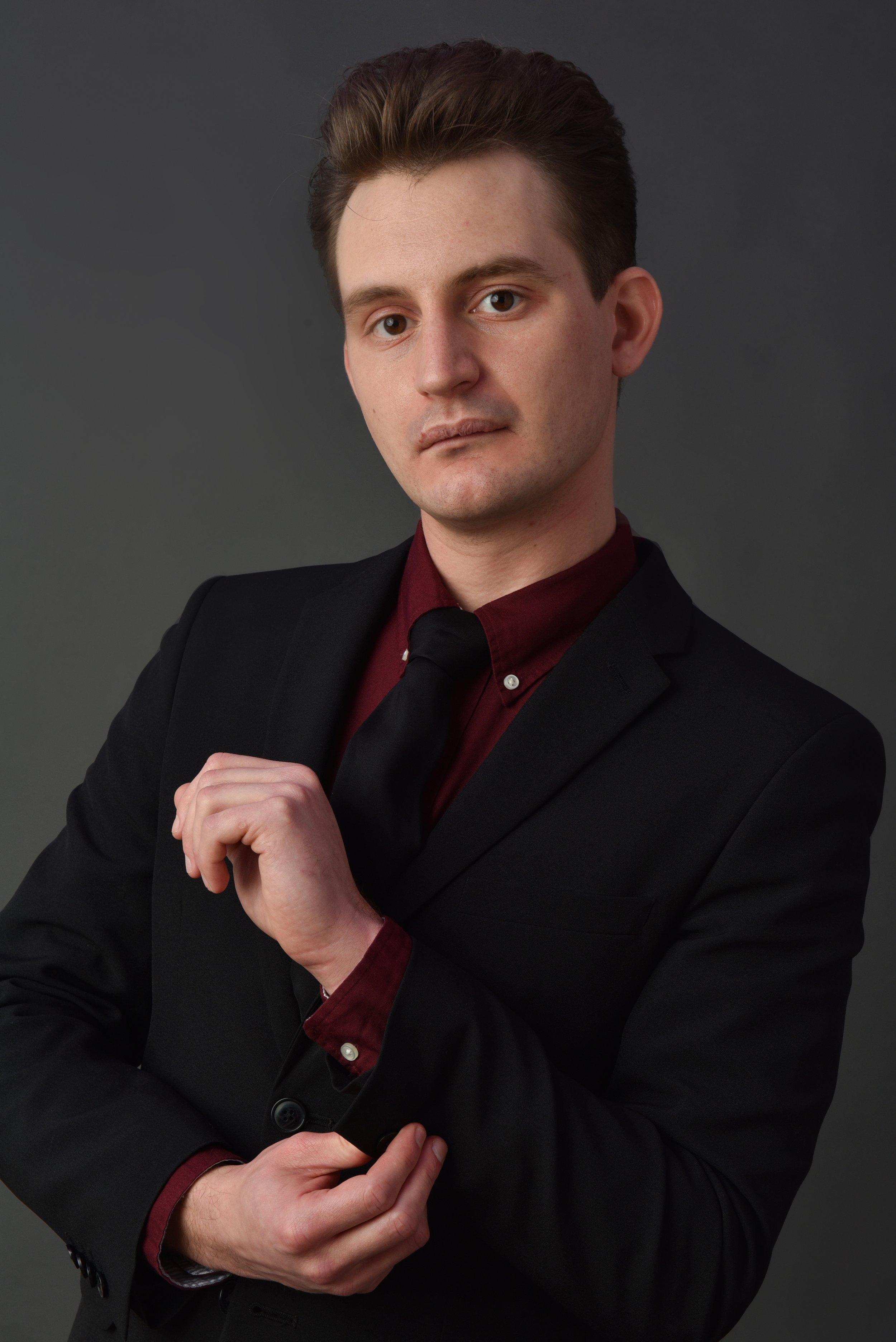A man in a black suit with a dark red shirt and black tie, looking serious, against a gray background.