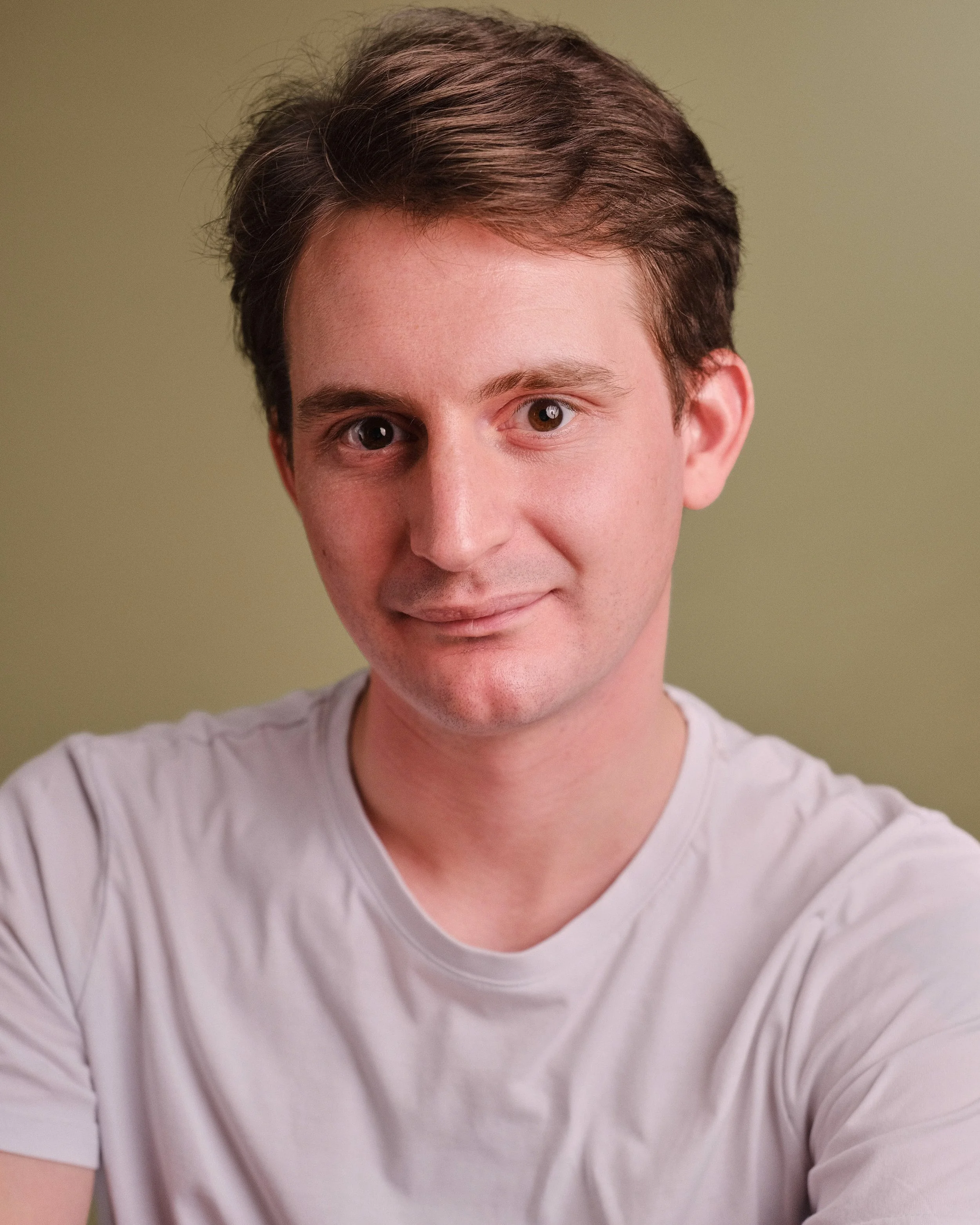 A young man with short brown hair and fair skin taking a selfie against a light green background, wearing a white t-shirt.