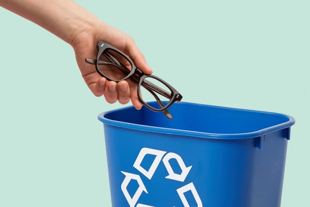 A hand is dropping a pair of black eyeglasses into a blue recycling bin with a white recycling symbol, against a light green background.