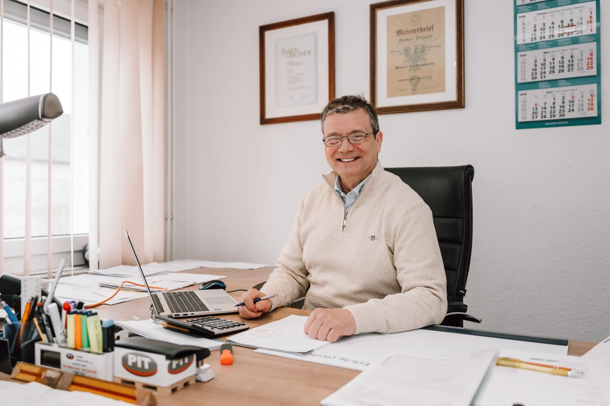 Ein lachender Mann sitzt an einem Büro-Schreibtisch mit Laptop, Papieren und Bürobedarf. Hinter ihm an der Wand hängen drei gerahmte Urkunden und drei Wandkalender. Es ist ein helles Büro mit Sonnenlicht, das durch ein Fenster scheint.