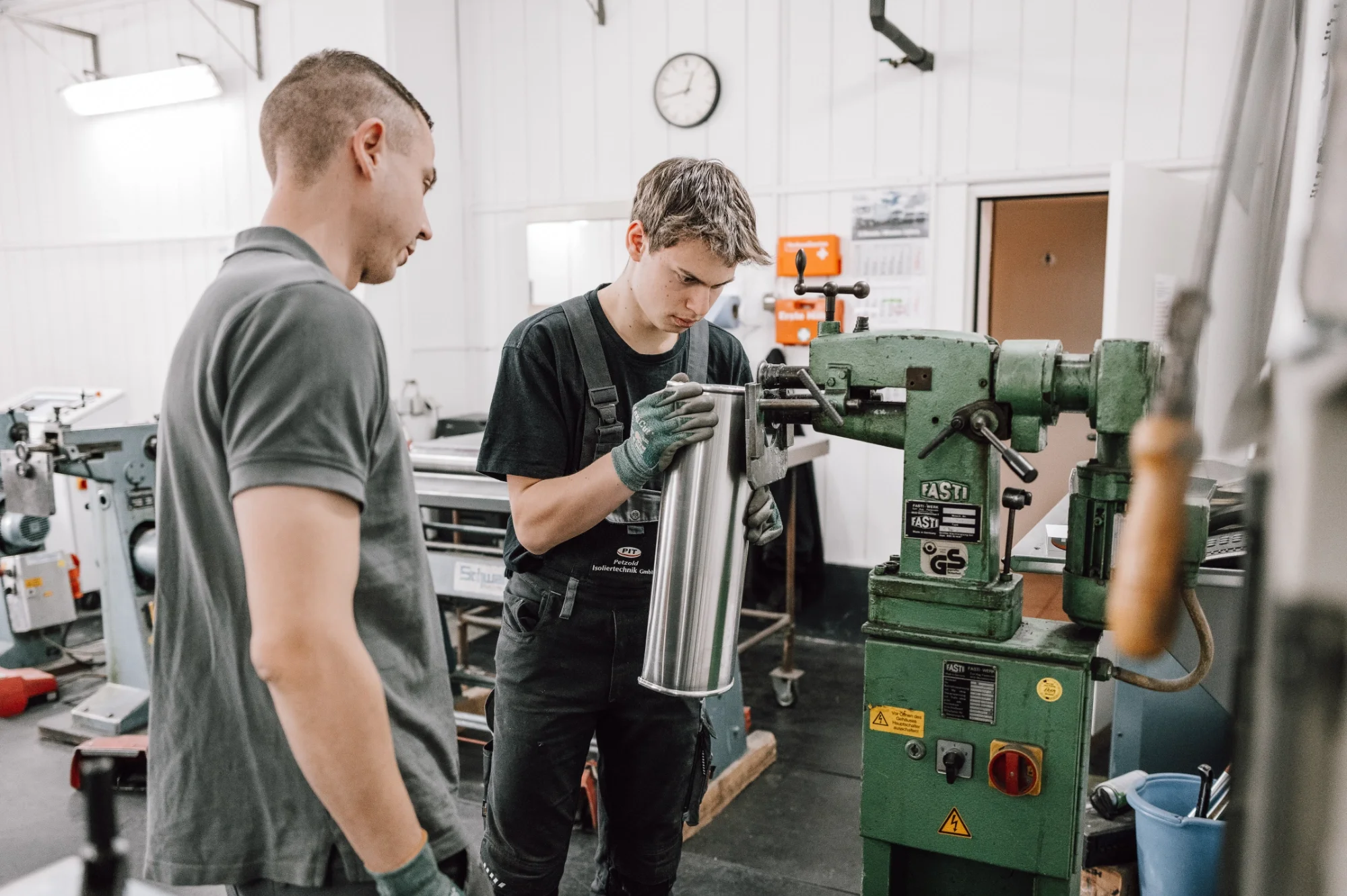 Zwei junge Männer arbeiten in einer Werkstatt mit einer grünen Maschine, einer misst einen Metallzylinder, der andere beobachtet.