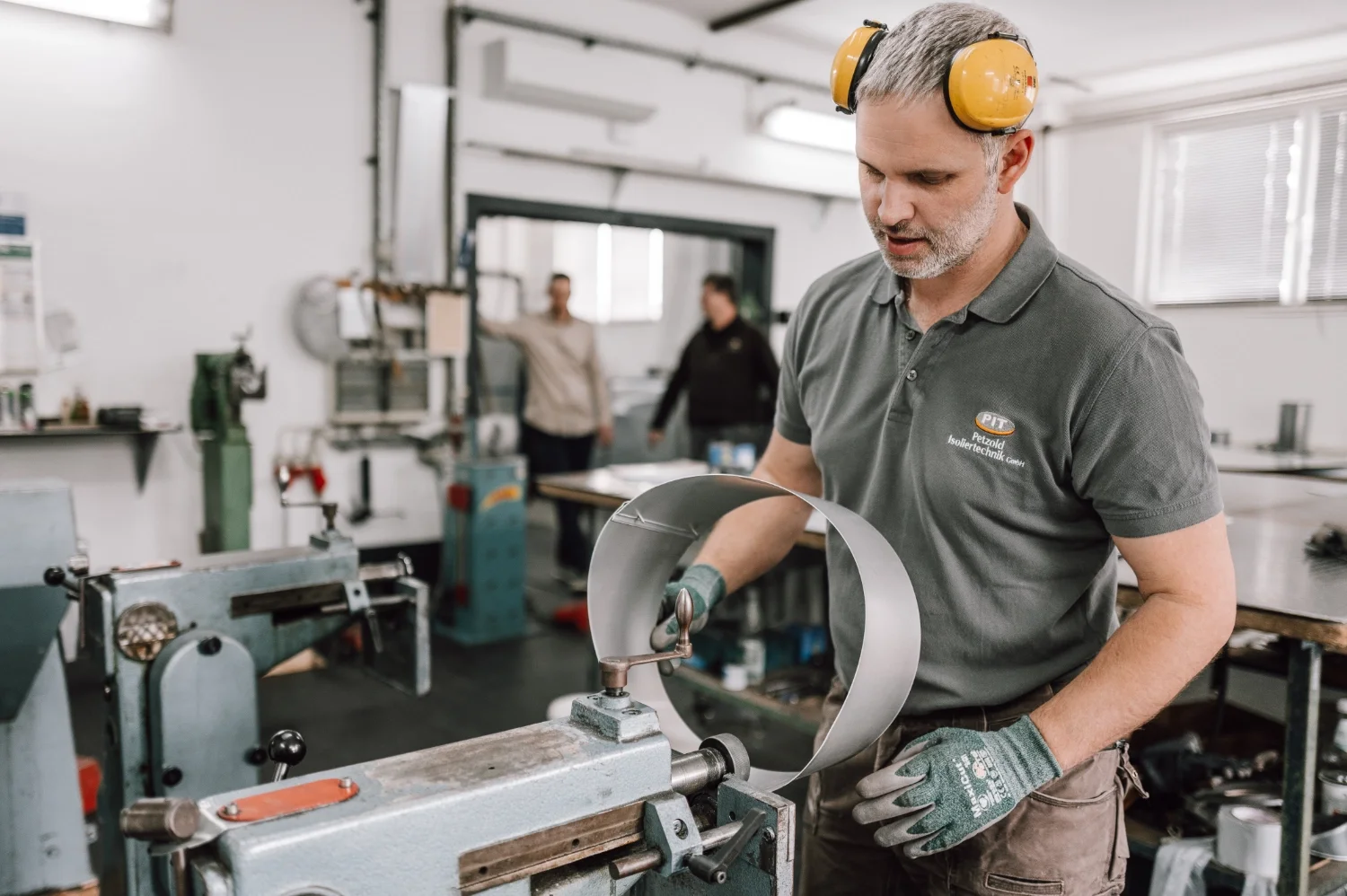 Ein Mann mit grauem Haar trägt Gehörschutz und arbeitet an einer Metallspule in einer Werkstatt, während zwei weitere Personen im Hintergrund stehen.