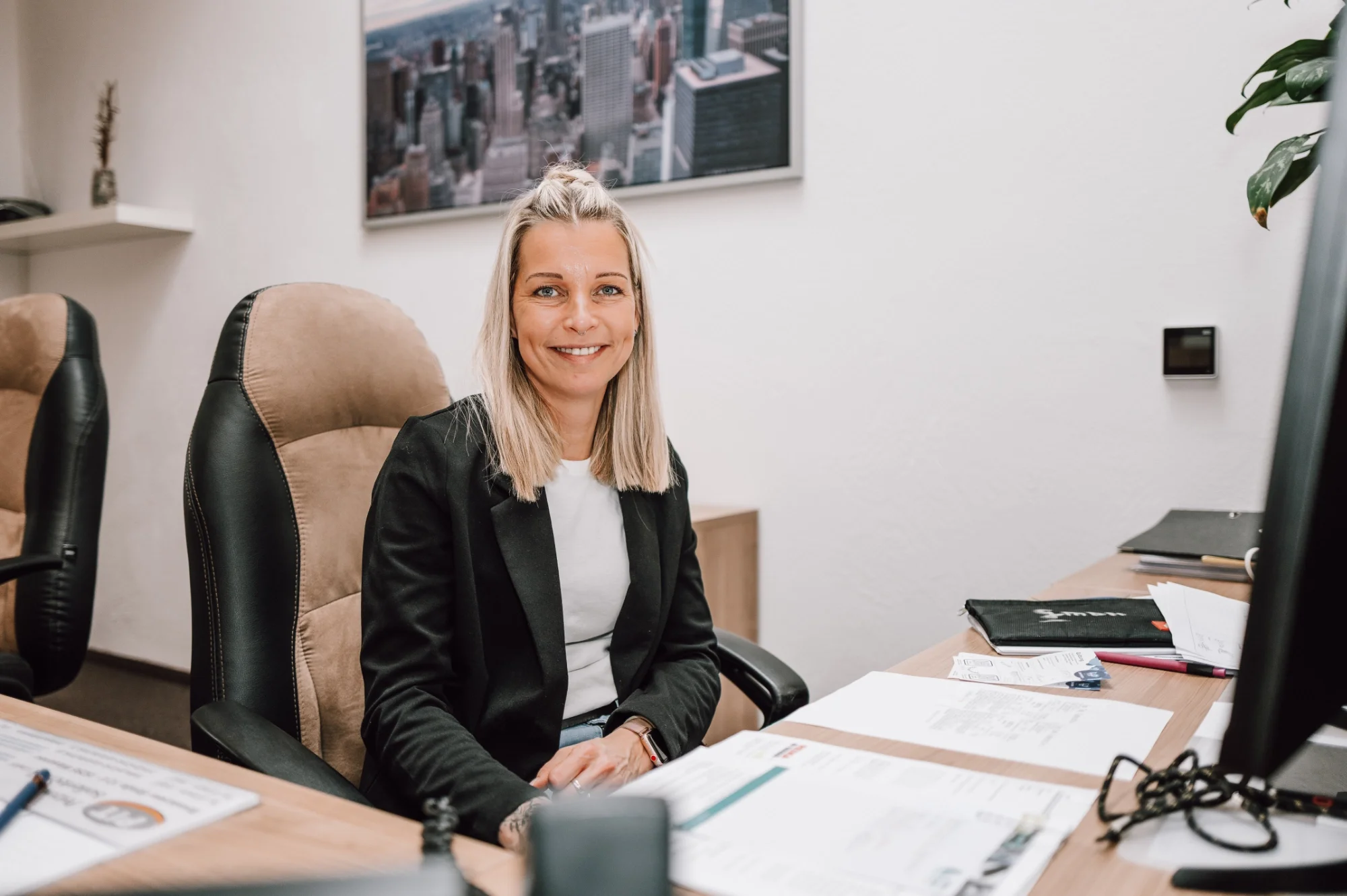 Eine lächelnde Frau mit blonden Haaren sitzt an einem Schreibtisch in einem Büro, umgeben von Papieren, Stiften und einem Aktenordner. Hinter ihr befindet sich eine Wand mit einem Stadtbildbild, und es sind einige Büroartikel auf einem Regal zu sehen.