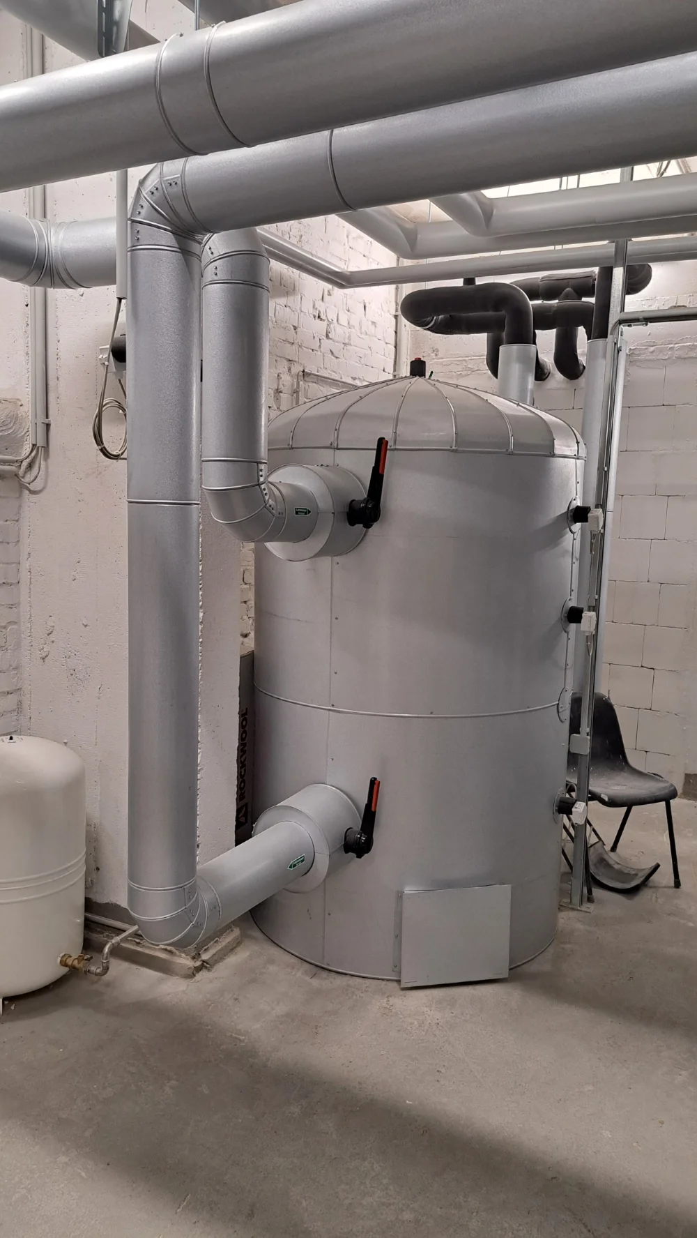 Industrieheizung oder Wärmetauscher in einem Kellerraum mit weißen Wänden und schwarzen Rohren.