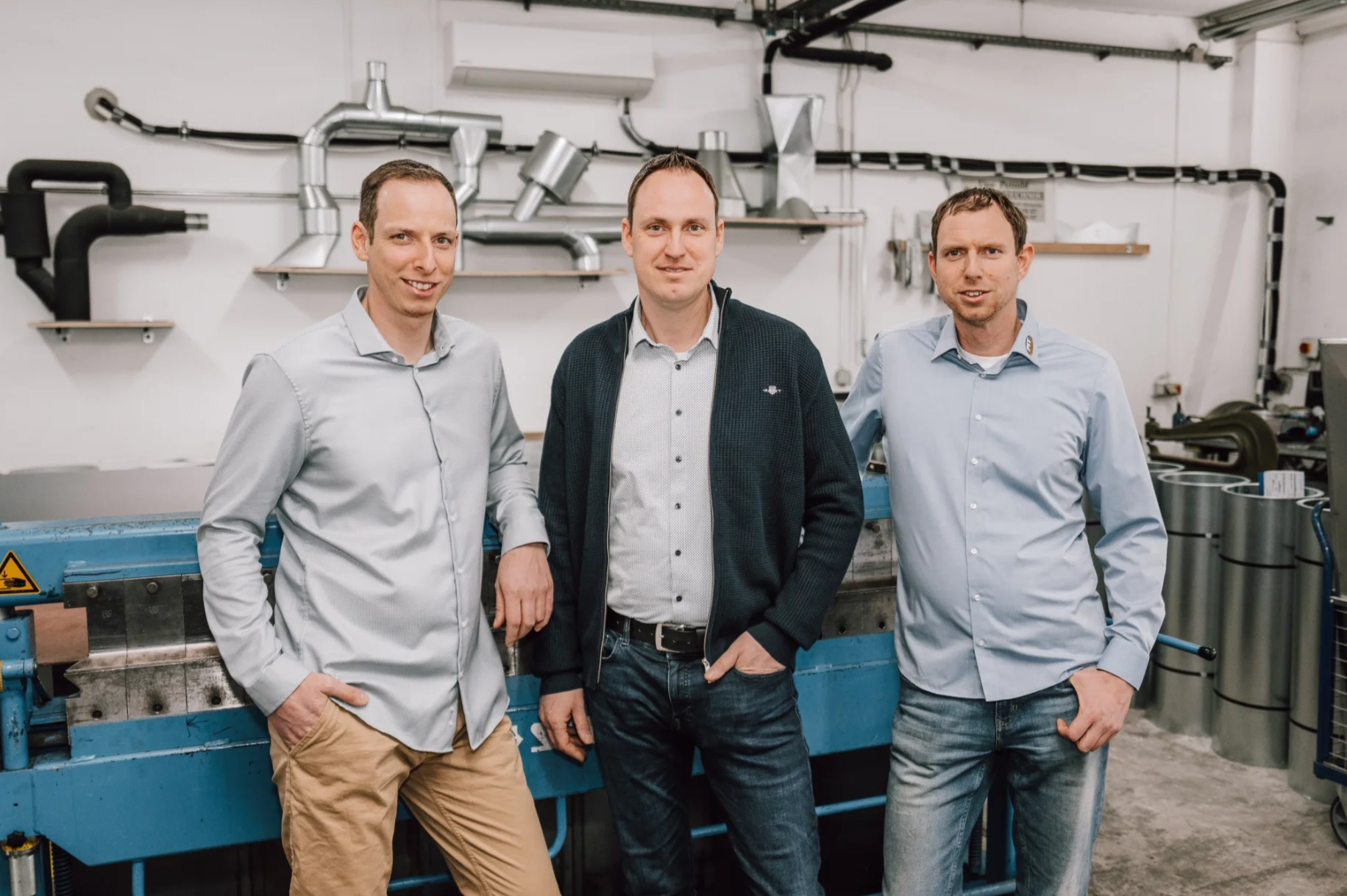 Drei Männer stehen in einer Werkstatt vor einer großen Maschine, die mit Rohren und technischen Einrichtungen verbunden ist.