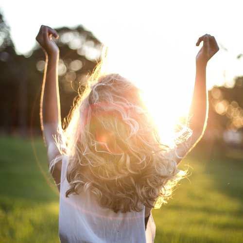 Ein Mädchen mit lockigem Haar, das nach vorne schaut und die Arme in die Luft hebt, bei Sonnenuntergang im Freien.