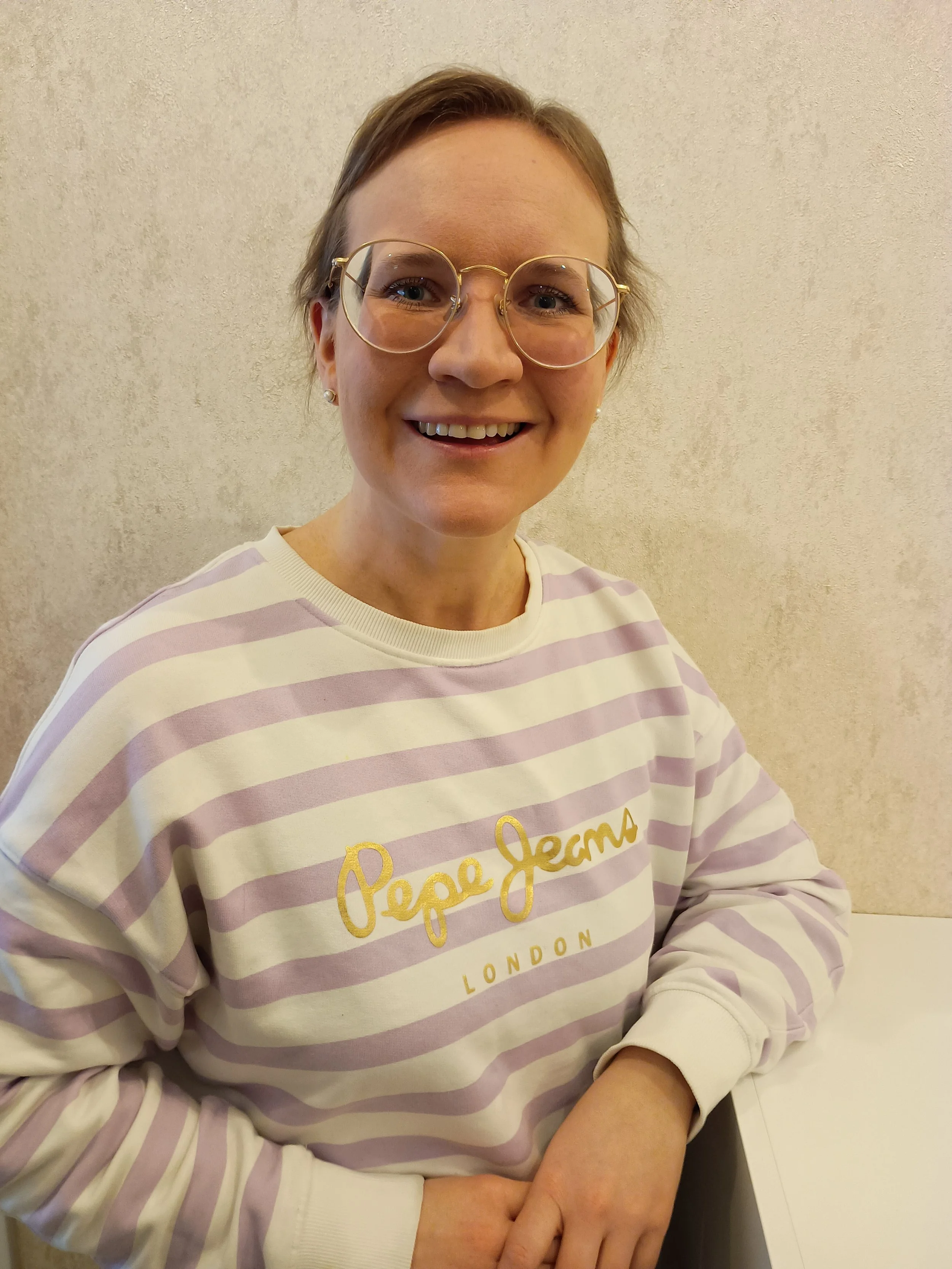 Portrait einer Frau mit Brille, kurz haar, lächelnd, trägt ein weiß-lila gestreiftes Sweatshirt mit goldener Schrift 'Pepe Jeans LONDON'.