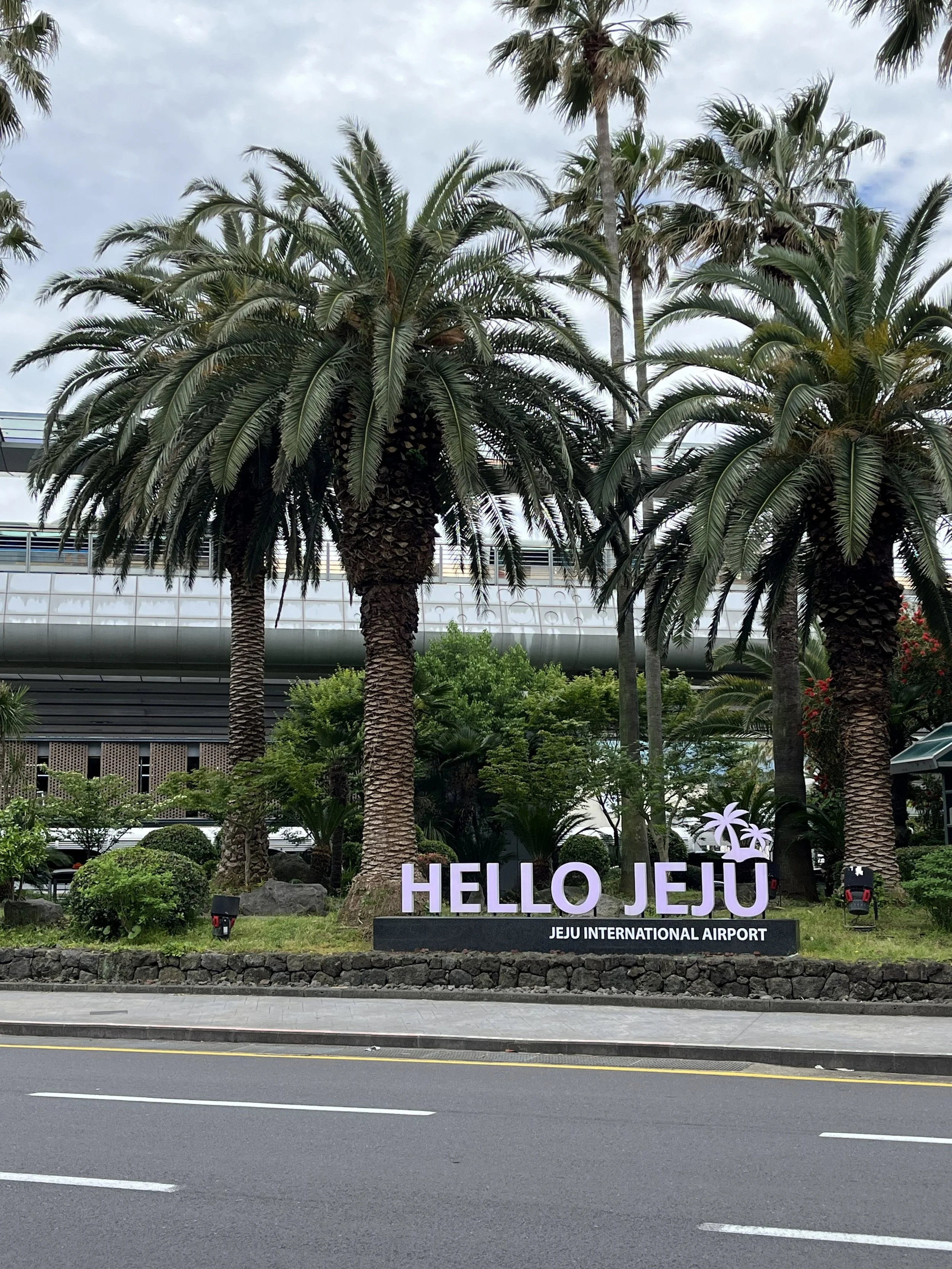Signalisation indiquant 'HELLO JEJU' en lettres violettes avec palmiers, devant un bâtiment moderne et des arbres à Jeju, aéroport international.