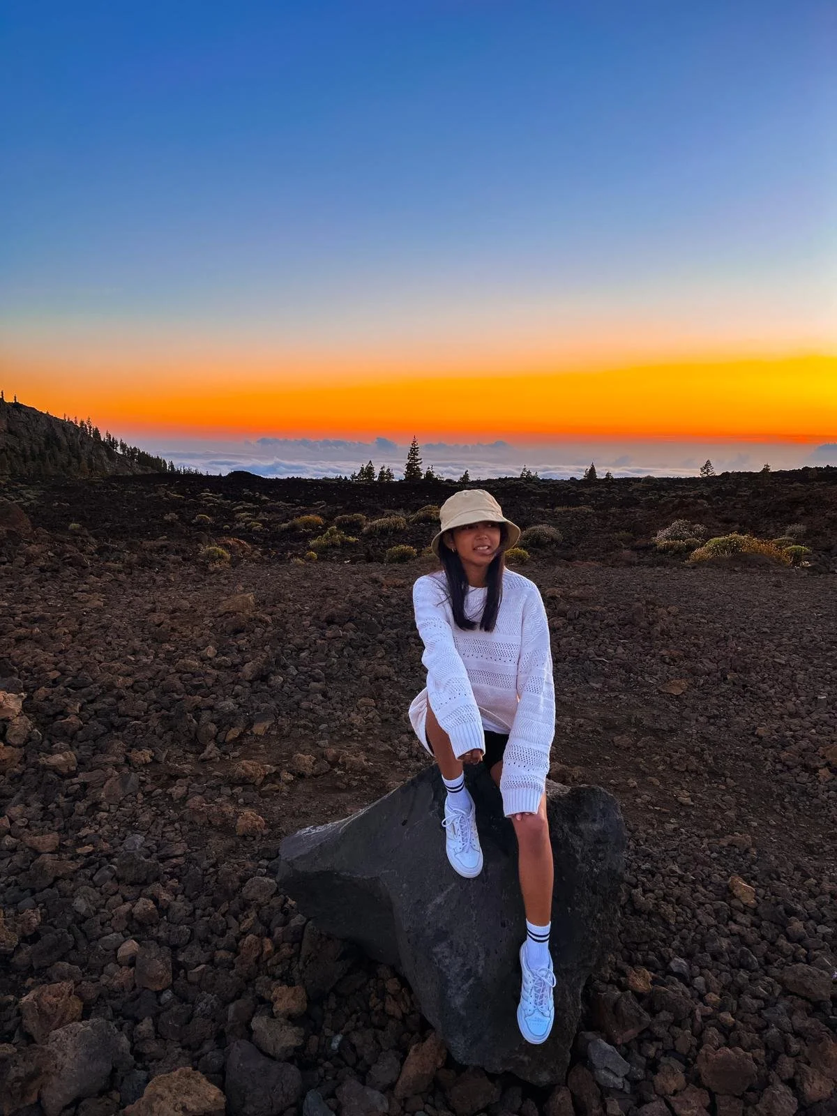 Jeune femme assise sur une roche dans un paysage volcanique au coucher du soleil, portant un chapeau beige, un pull blanc, des baskets blanches et des chaussettes rayées.