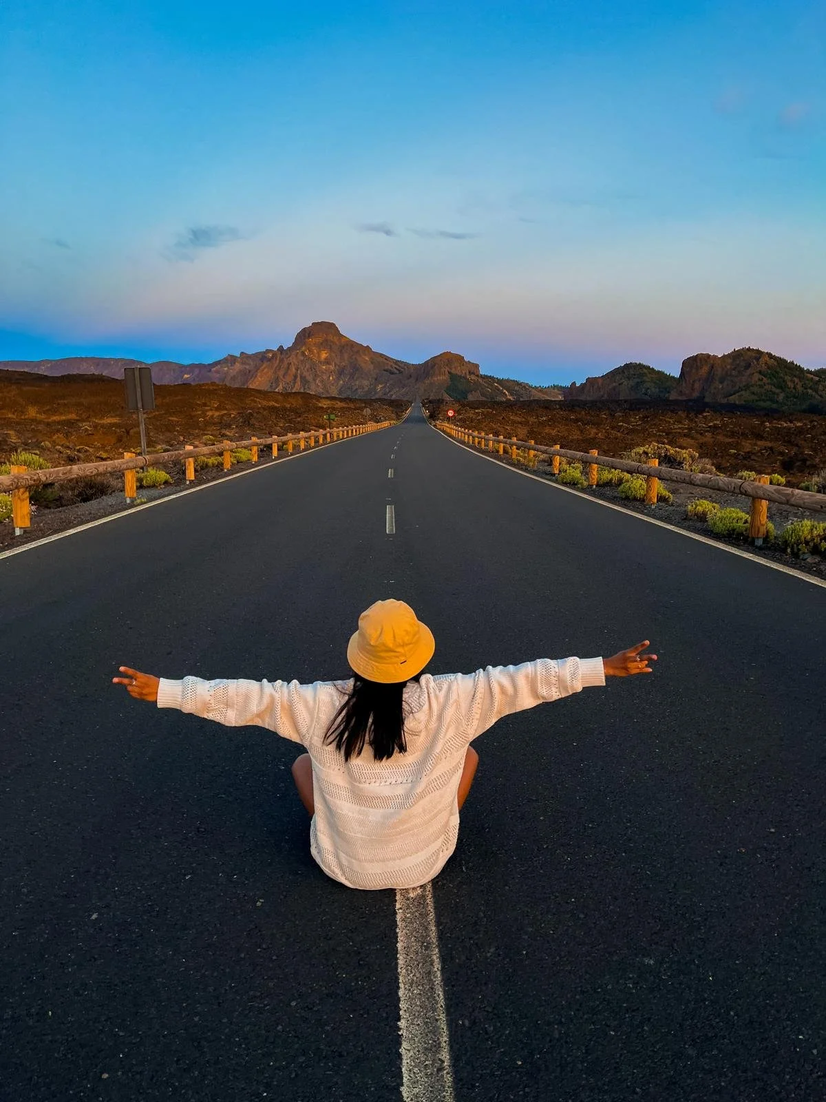 Une personne portant un chapeau jaune, assise au centre d'une route, avec les bras étendus, regardant vers l'horizon lors d'un coucher de soleil ou du lever du soleil, avec des montagnes en arrière-plan.