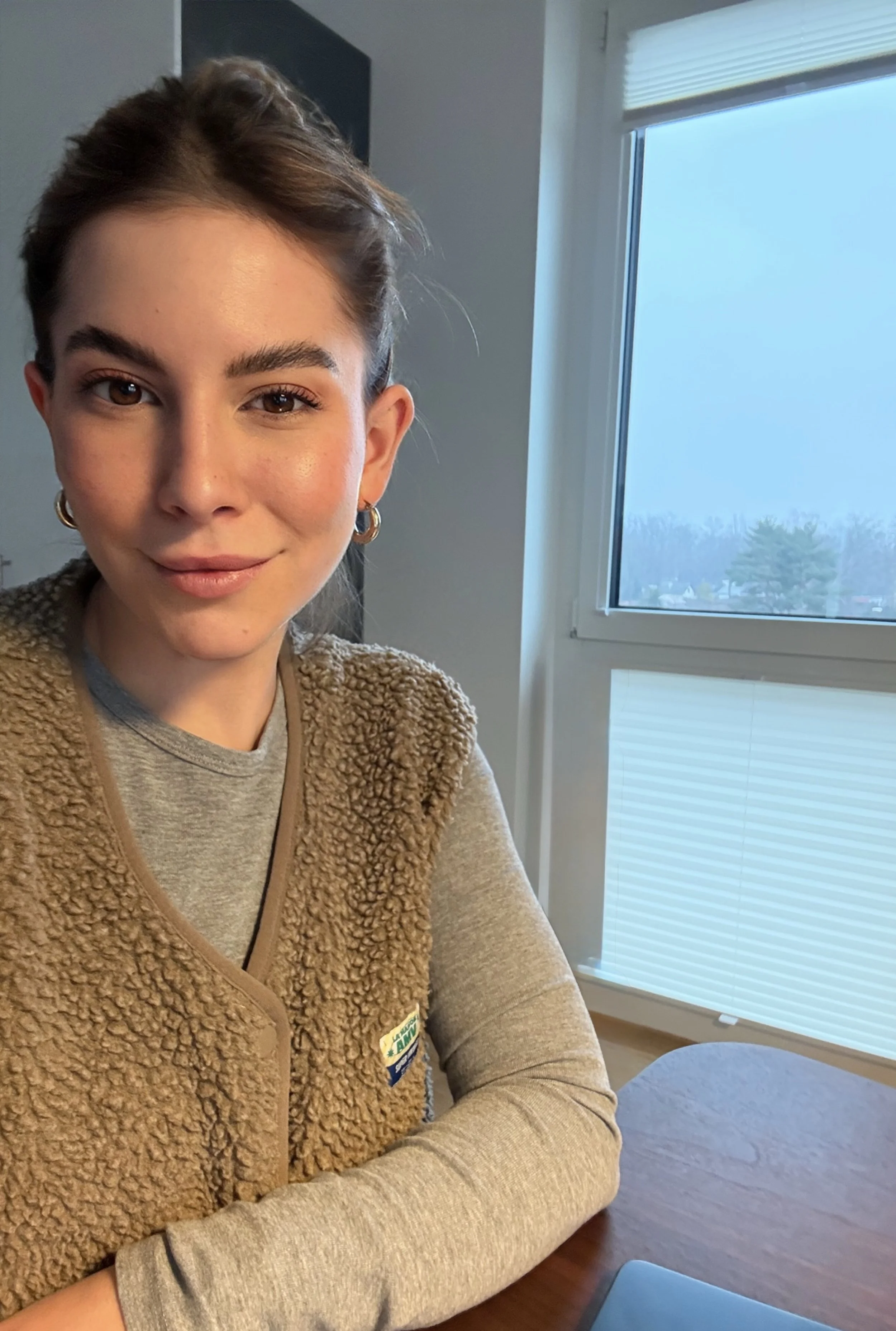 Junge Frau sitzt an einem Tisch neben einem großen Fenster mit Blick nach draußen, trägt eine braune Weste und einen grauen Pullover, lächelt leicht, mit Earcuffs.