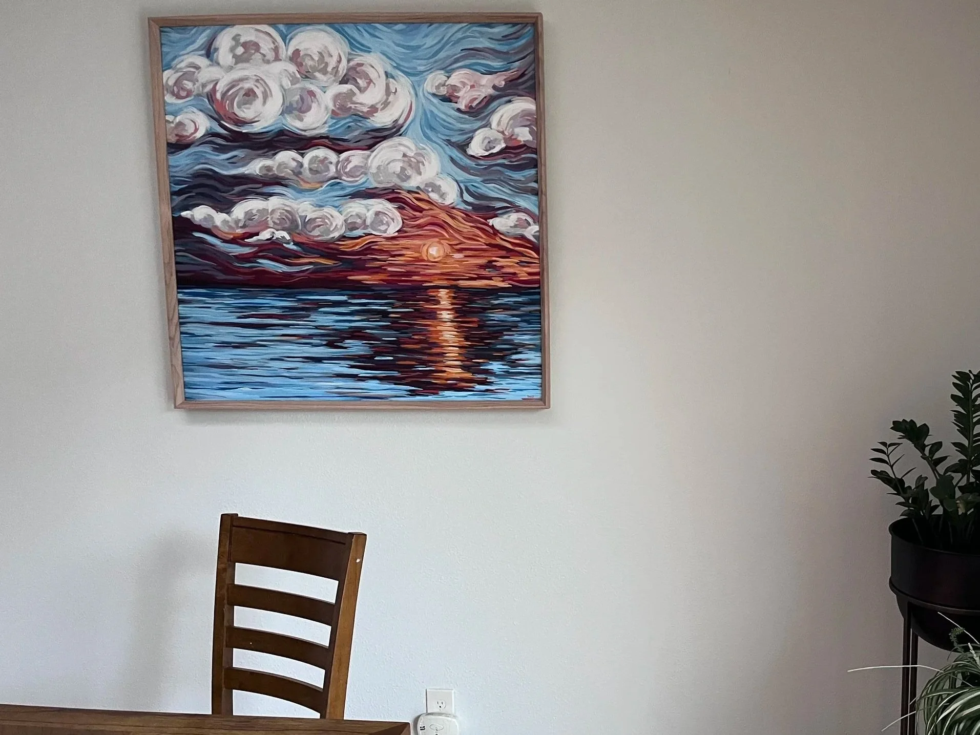 A framed painting of a sunset over water with clouds in a wooden frame hangs on a light-colored wall. There are two potted plants on black stands to the right, one taller with broad leaves and the other with long, narrow, green leaves with white edge