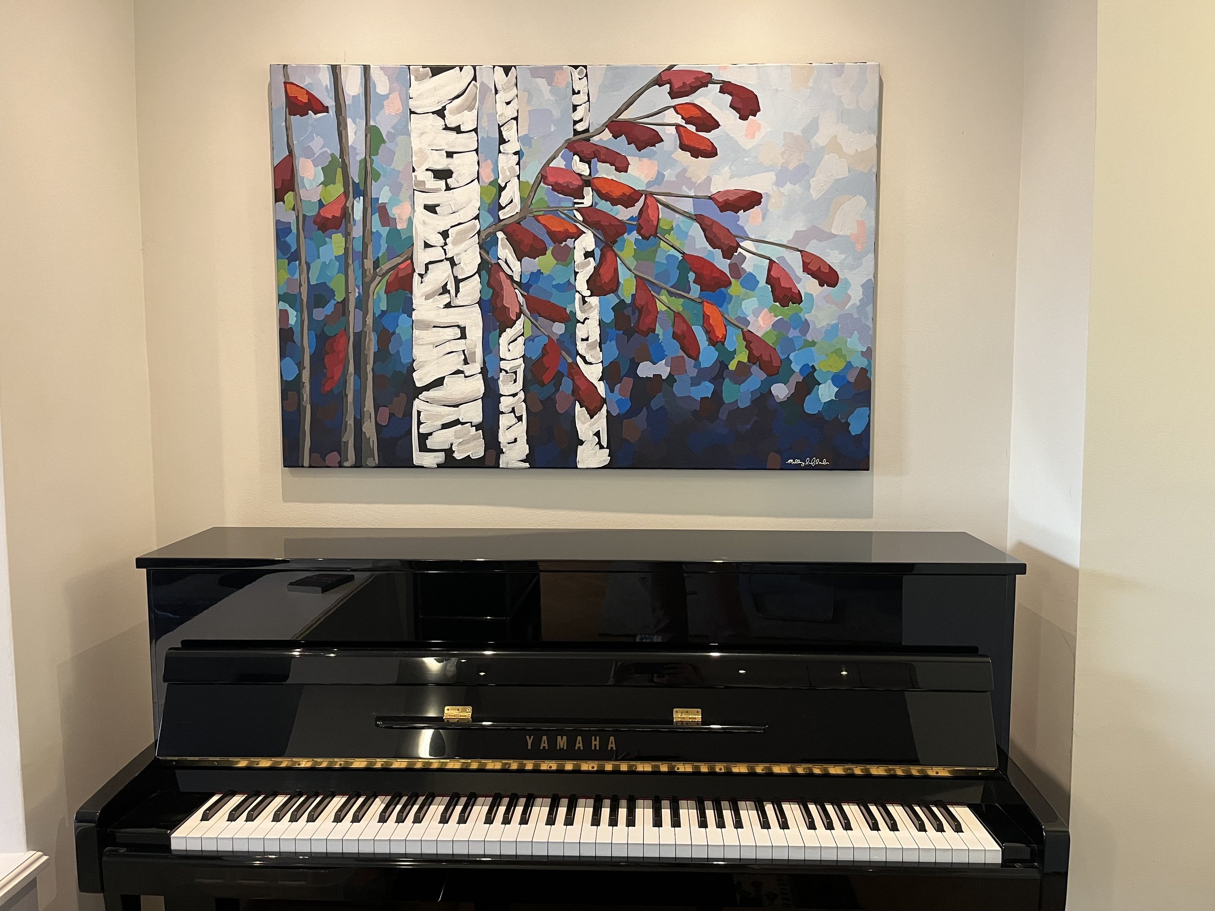 A black Yamaha upright piano in front of a wall with a colorful painting of white birch trees and red leaves.