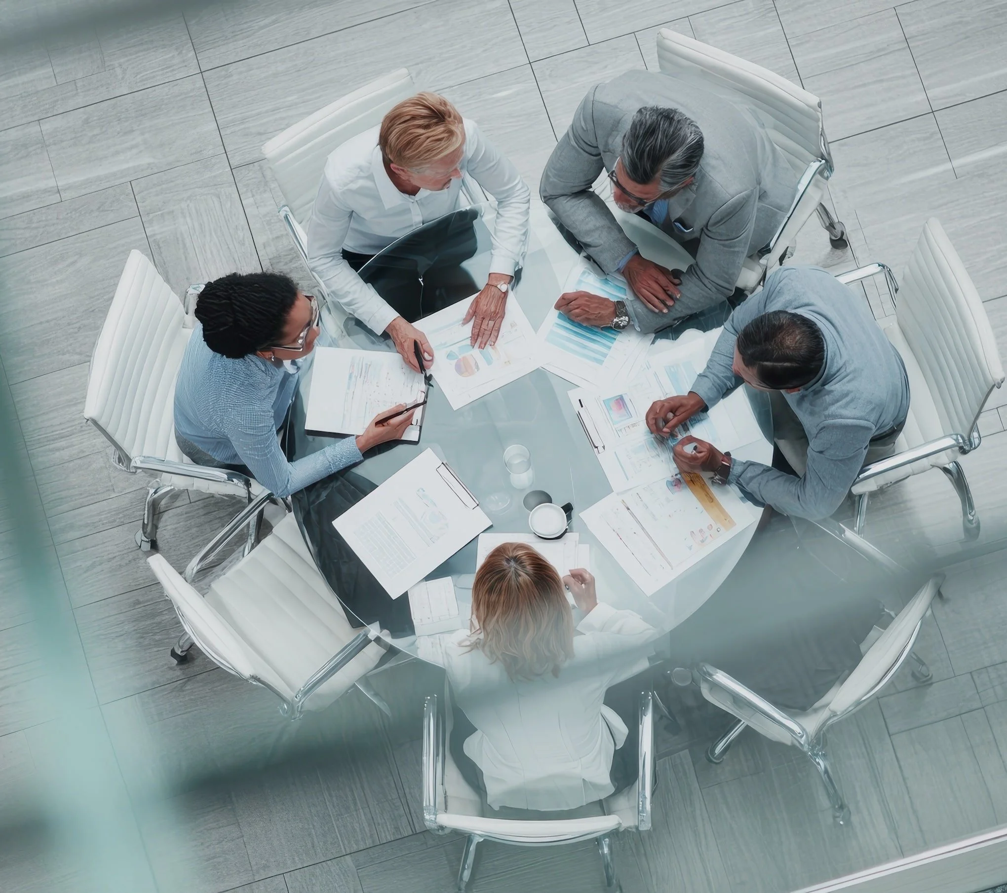 Top view of five business professionals sitting around a glass conference table, discussing financial reports and charts in a modern office.