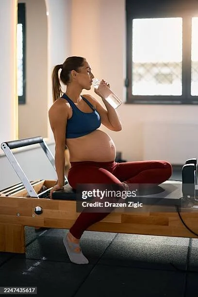 Pregnant woman in workout clothes drinking water while sitting on a Pilates reformer machine in a gym.