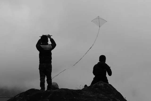 Zwei Personen als Silhouetten auf einem Felsen im Nebel, eine lässt einen Drachen steigen, schwarz-weiß