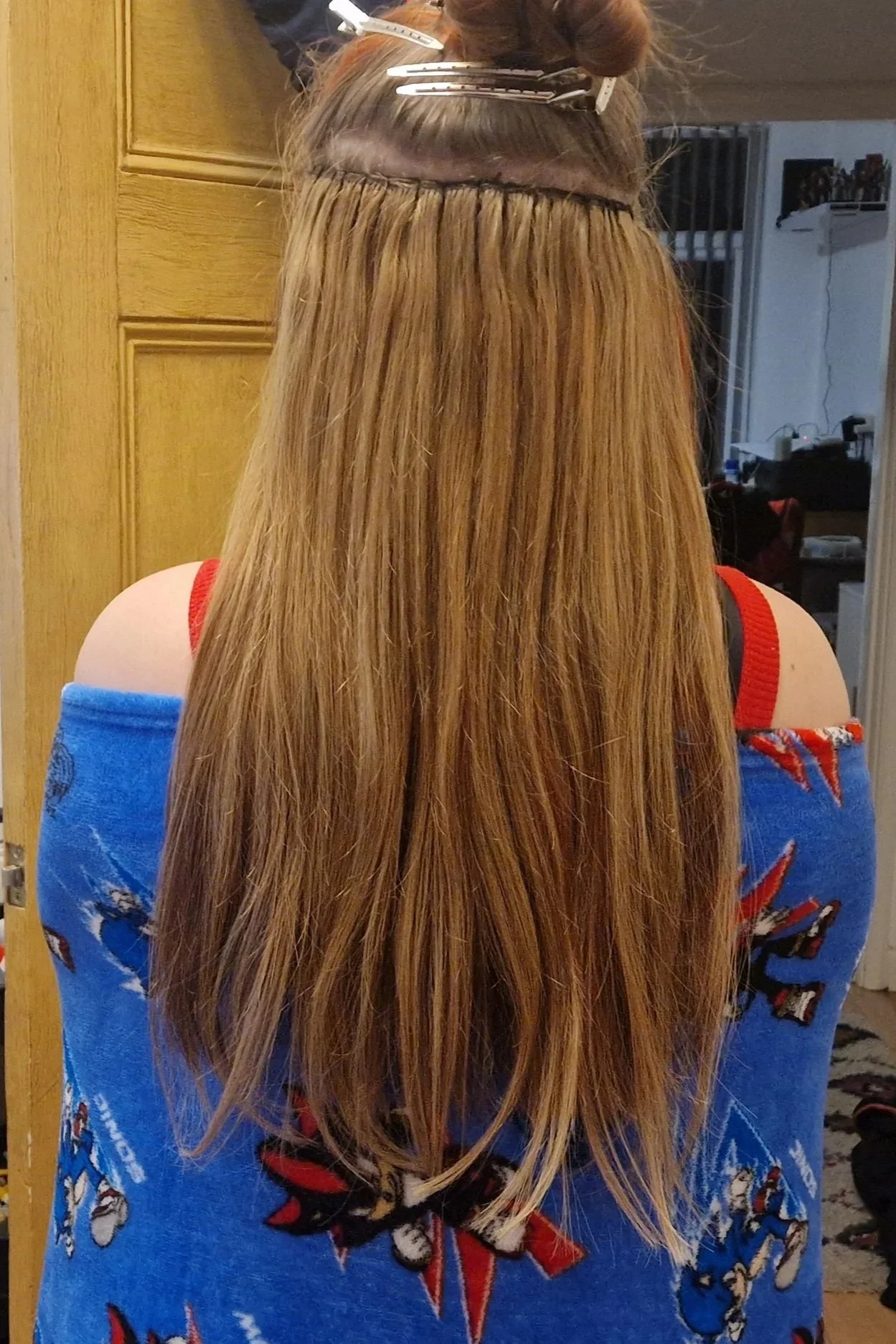 View of a woman with long, straight, blonde hair from behind, sitting in a chair with her hair in the process of being styled. The woman is wearing a blue off-shoulder top with red and white floral patterns, and her hair is sectioned and clipped at t