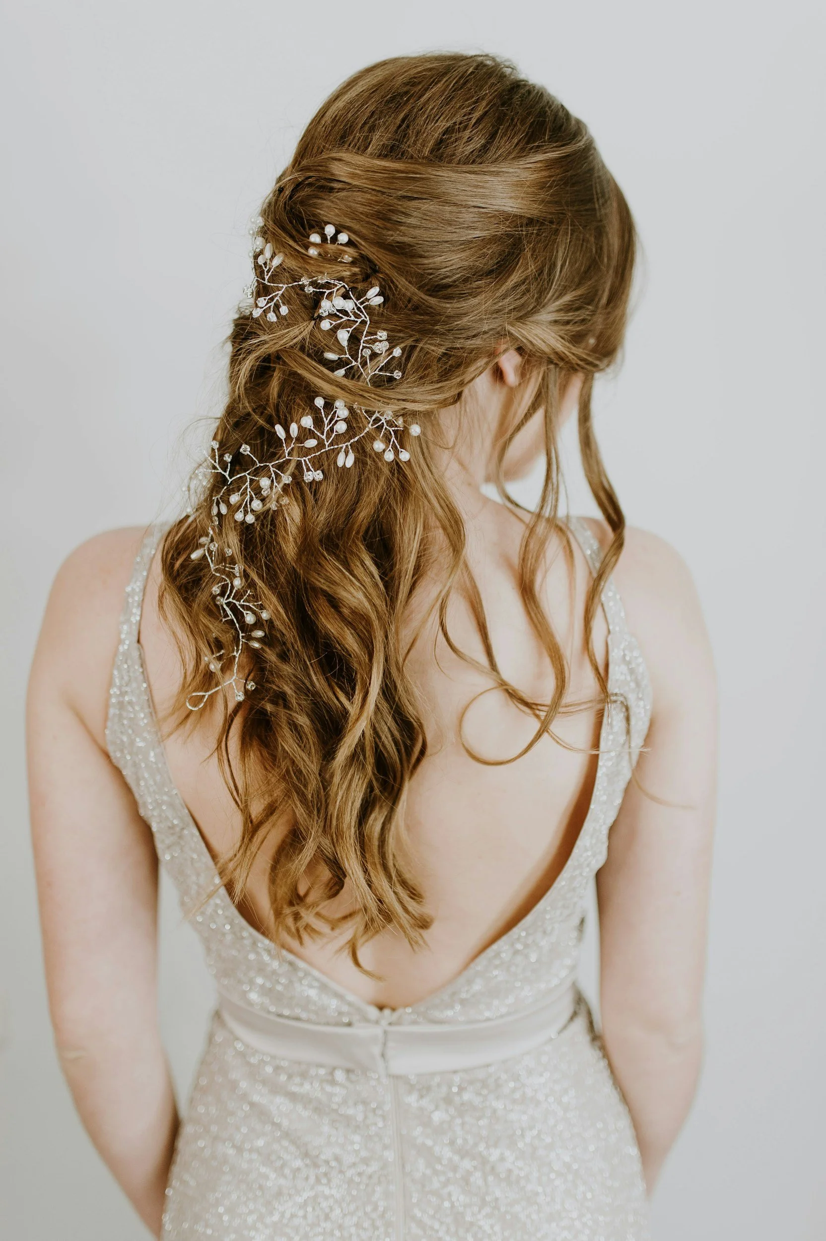 A woman with long, wavy, auburn hair styled with a delicate white floral hairpiece, wearing a sleeveless, V-neck, glittering wedding gown with intricate beadwork, standing against a plain white background.