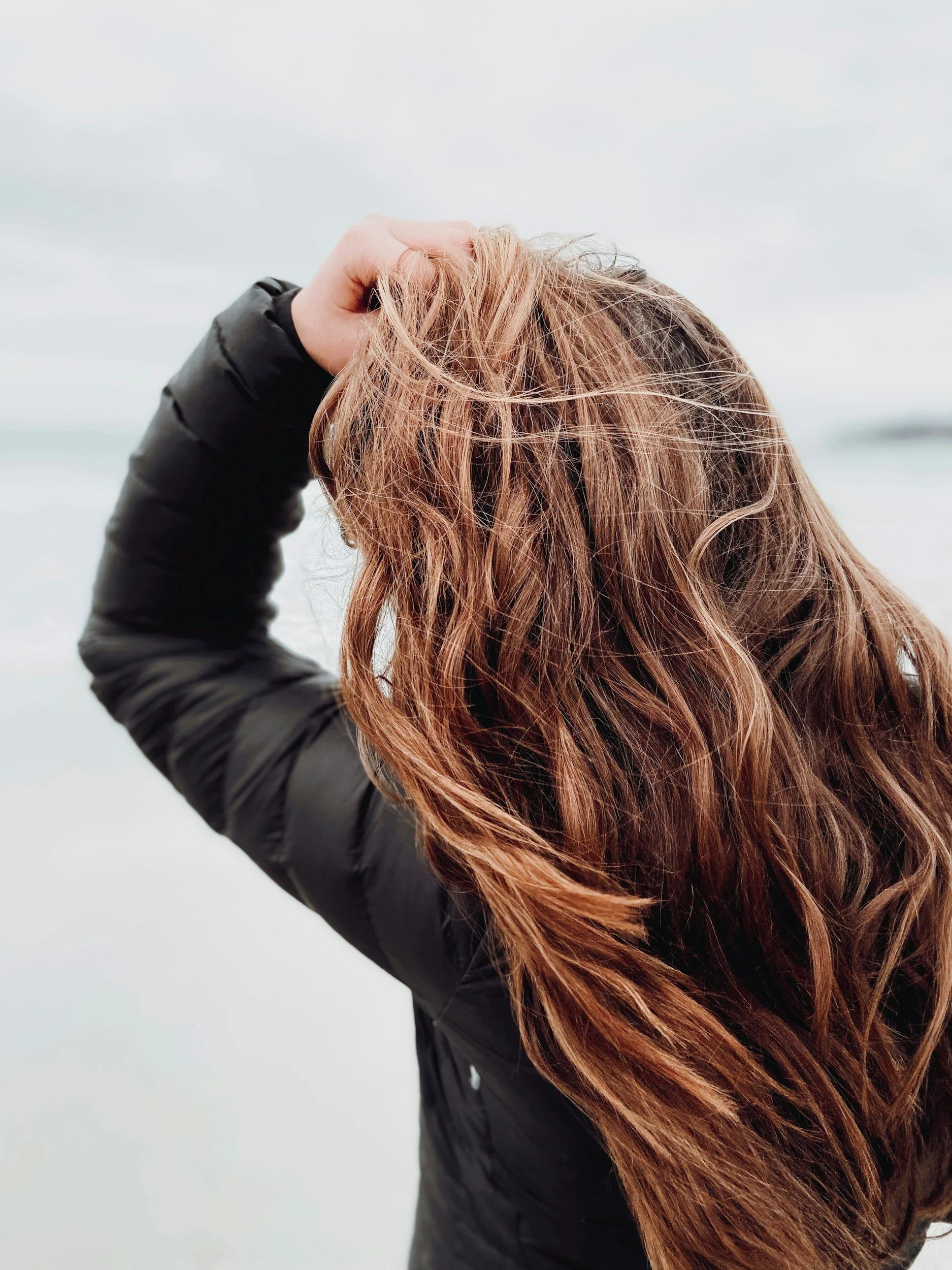 A person with long, wavy reddish-brown hair, wearing a black puffer jacket, holds their hand up to their head, as if they are playing with or adjusting their hair. The background appears to be a cloudy sky.