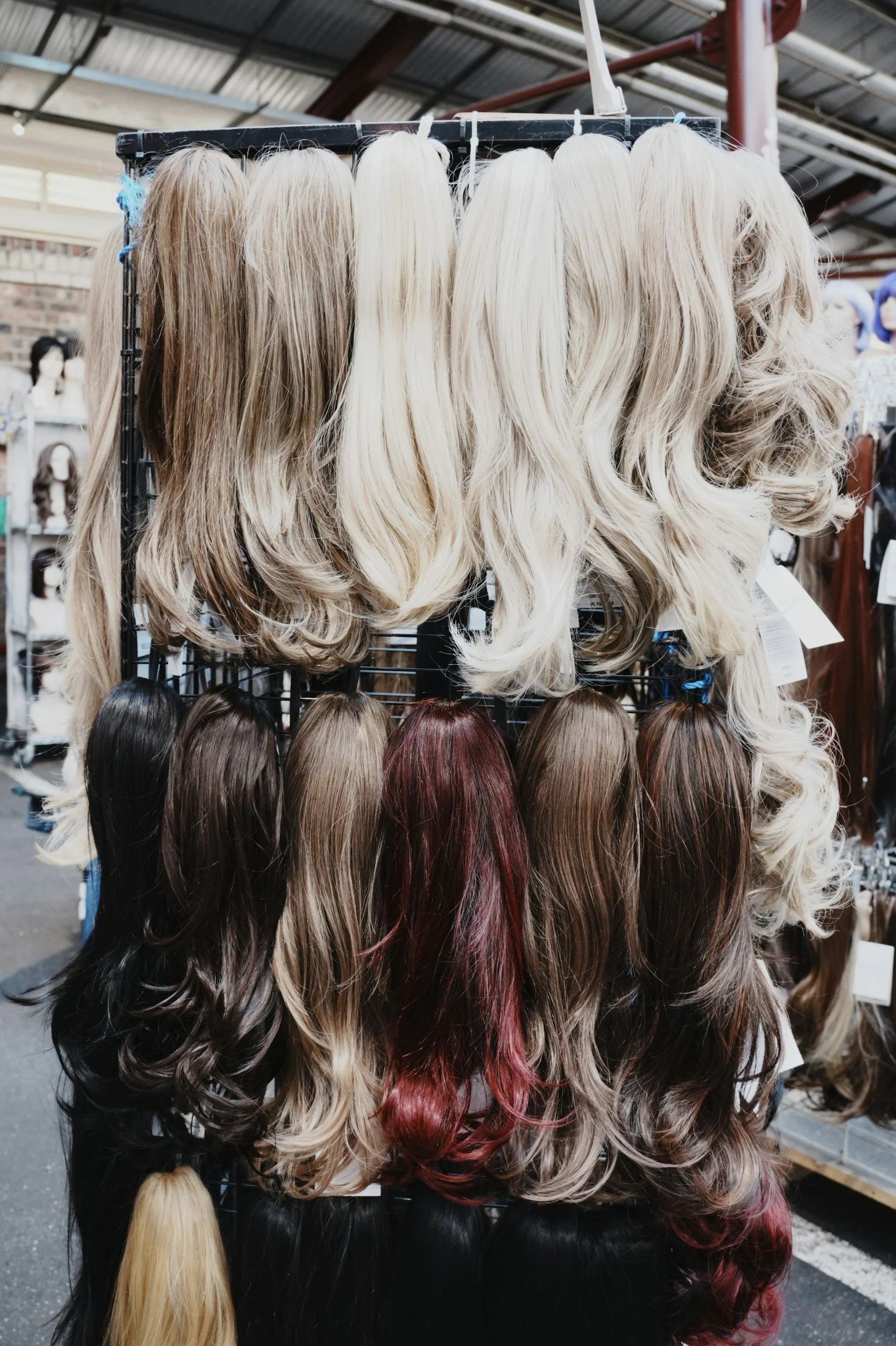 Display of various mannequin wigs in different shades of blonde, brunette, and red, hanging on a rack inside a warehouse.