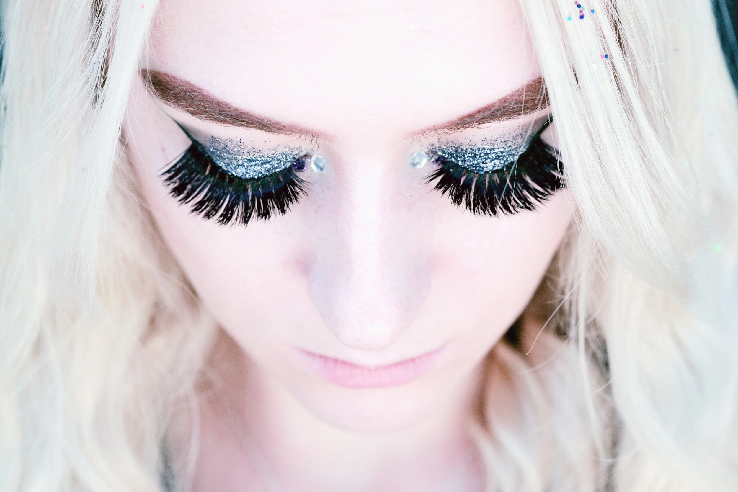 Close-up of a woman with platinum blonde hair and dramatic makeup, wearing silver and black glittery eyeshadow, rhinestones near her eyes, and long false eyelashes.