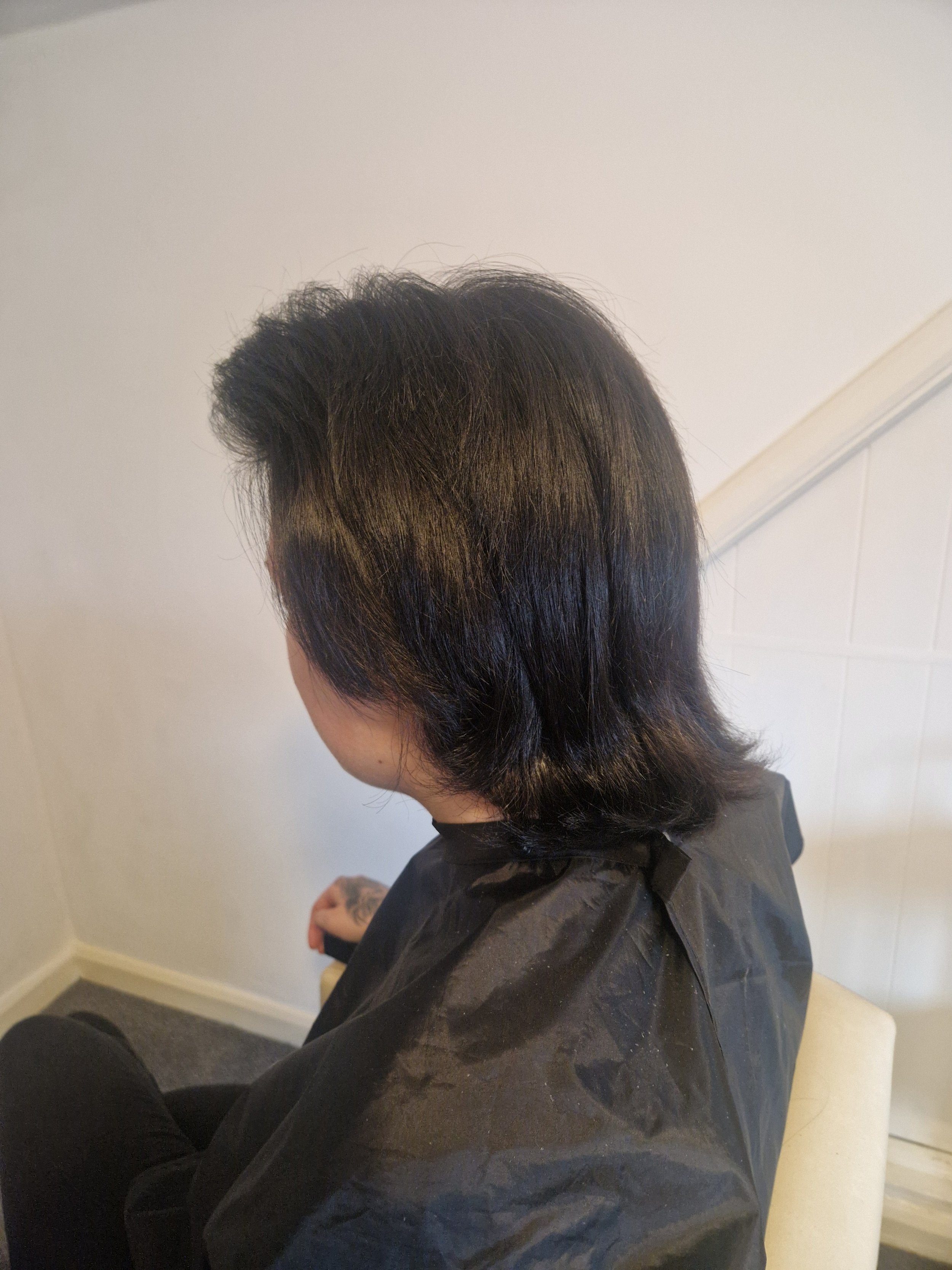 Person sitting in a salon chair with freshly styled, dark, layered hair, wearing a black salon cape, in a room with white walls and wall paneling.