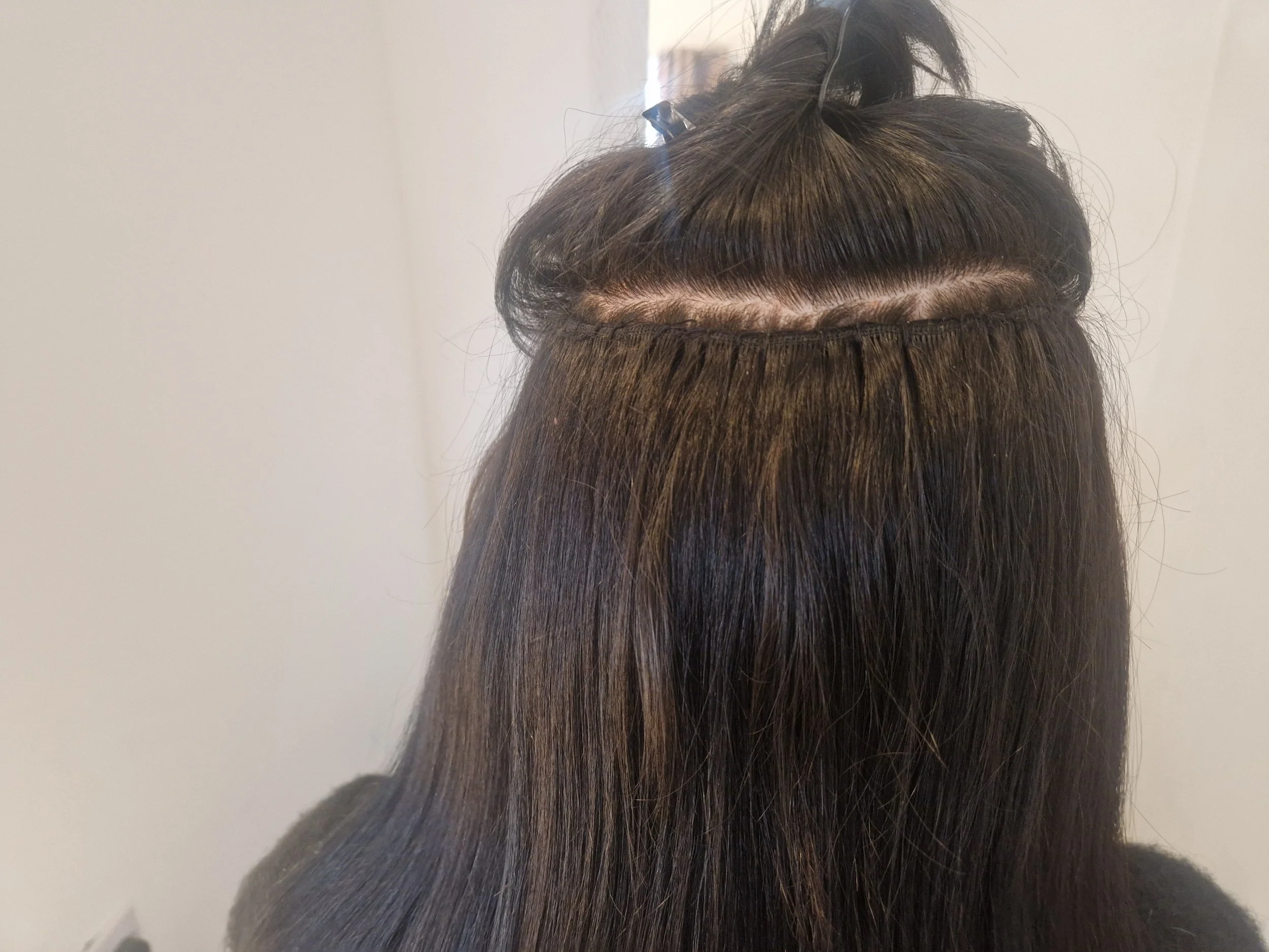 Close-up of a person's head showing the back of their long, straight, brown hair with a section that has been prepared for a hair extension or weaving process, secured with a small clip.