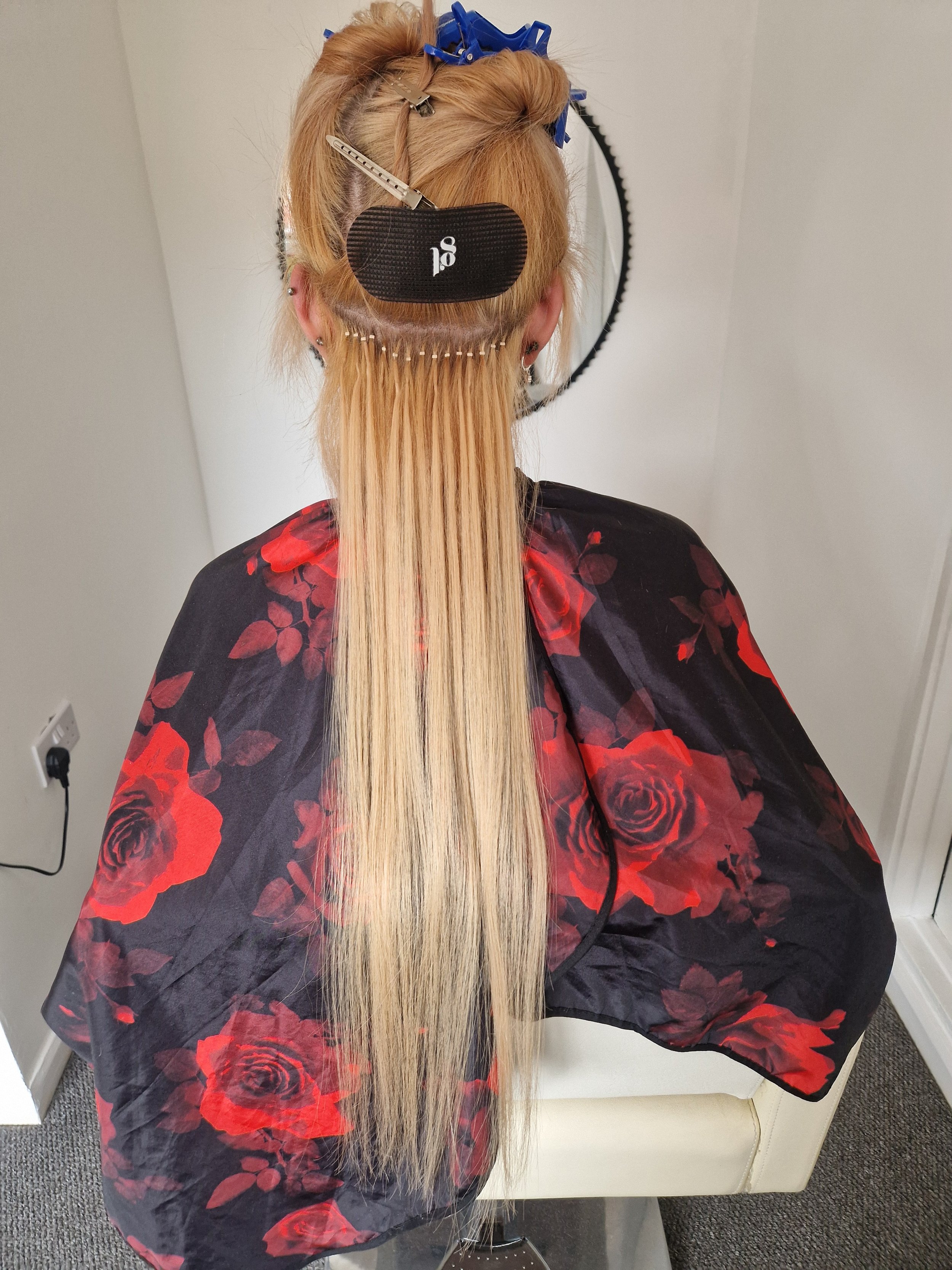 A woman with long blonde hair getting a hair treatment with rollers and clips, wearing a hair covering cap, seated in a salon with a white couch and gray carpet.