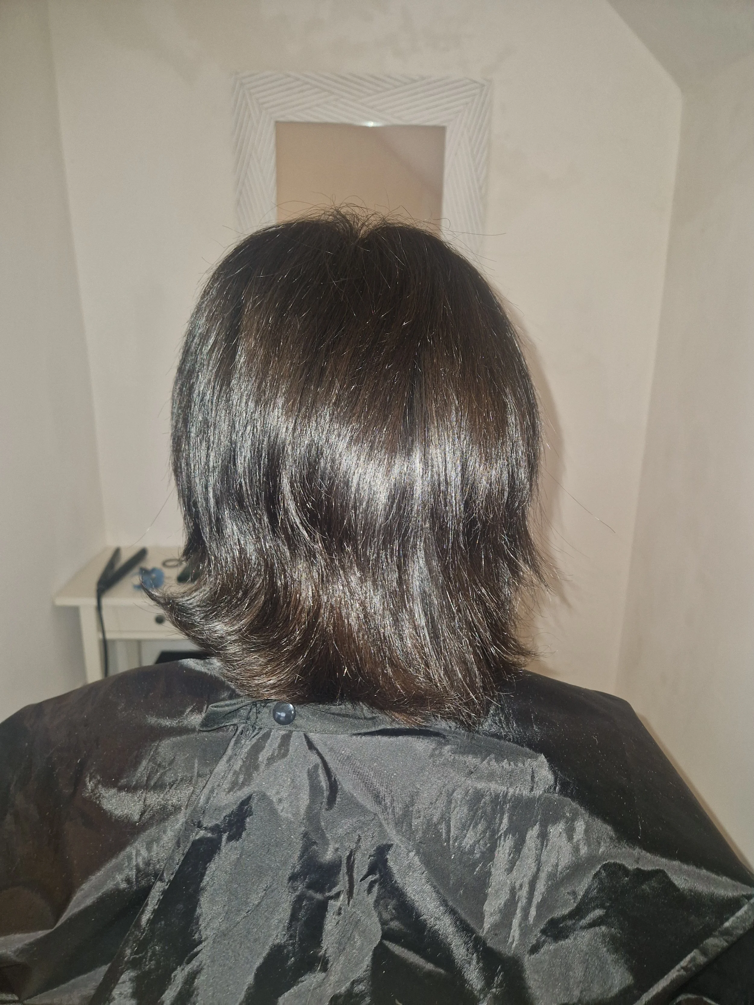 Back of a person's head with shiny, shoulder-length dark hair, wearing a black cape, in a room with a mirror and a white table with hair styling tools.