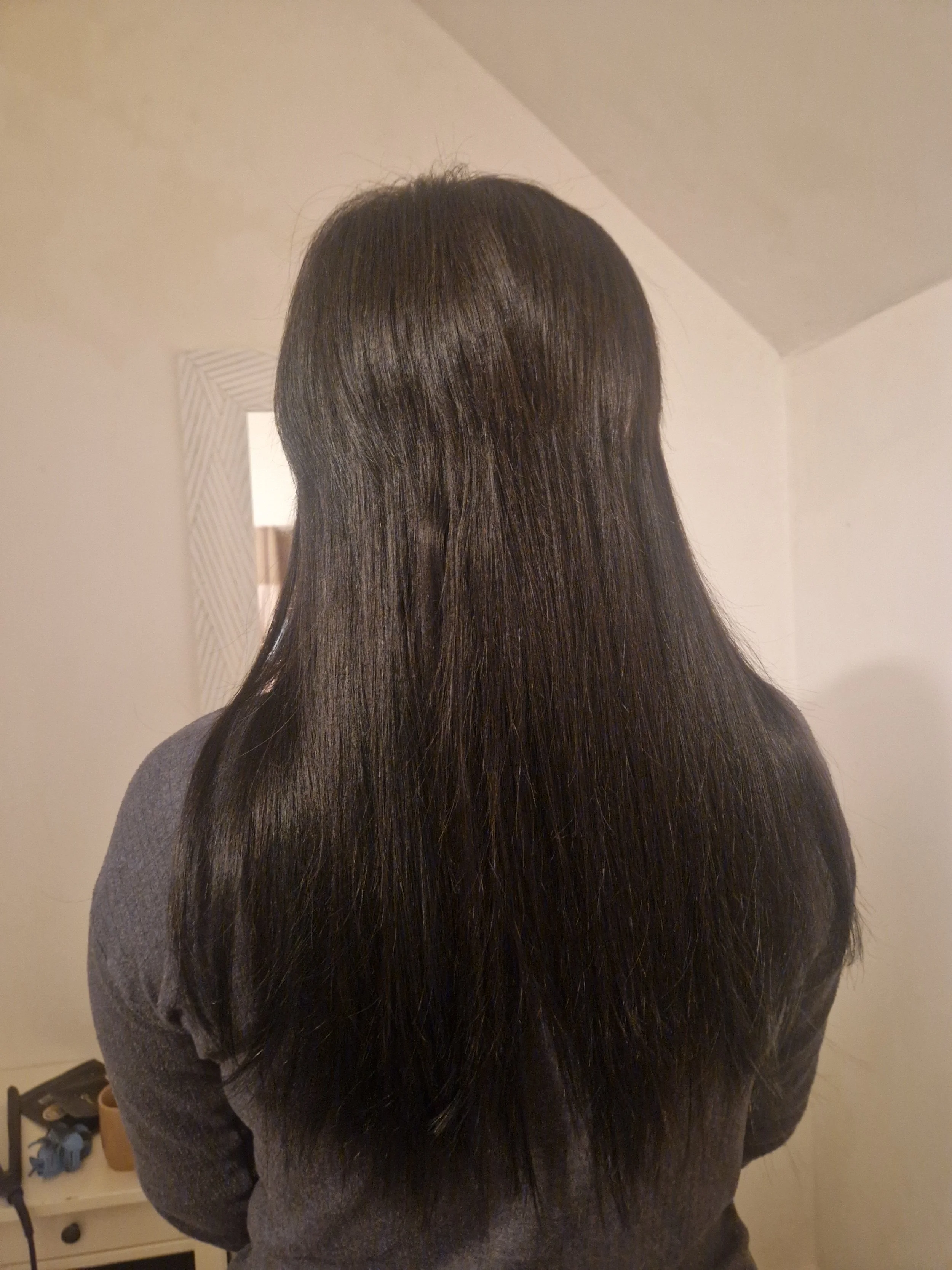 Back view of a person with long, straight, shiny black hair, wearing a gray sweater, standing in front of a mirror in a room with beige walls.