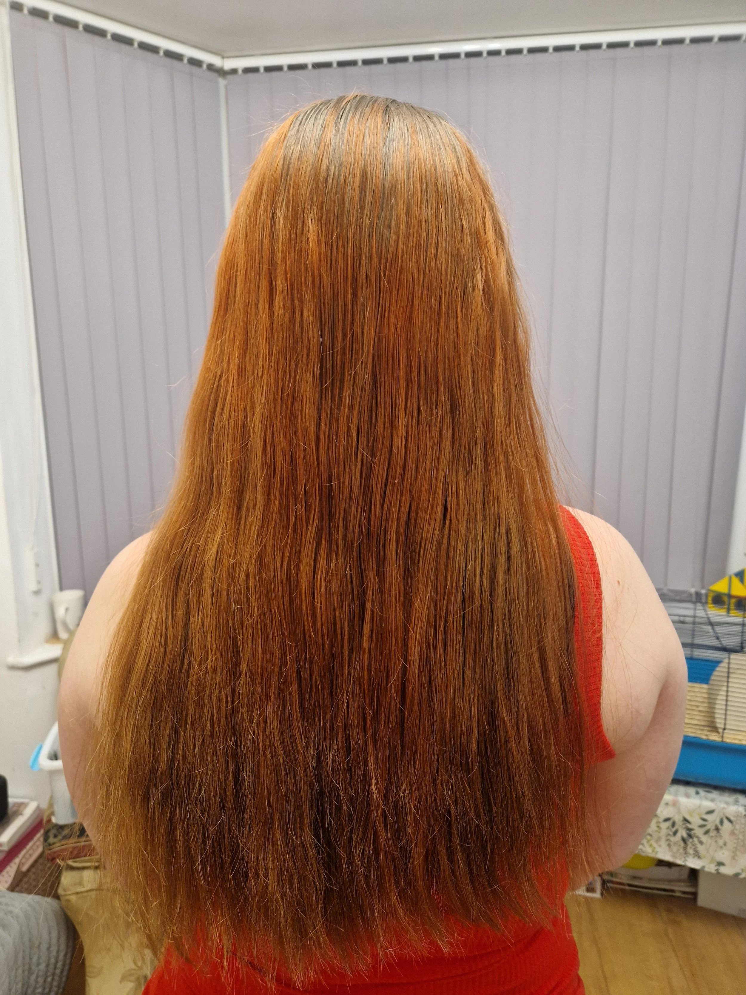 Back view of a person with long, straight, reddish hair, wearing a red sleeveless top, standing indoors with a gray wall and a wire cage in the background.