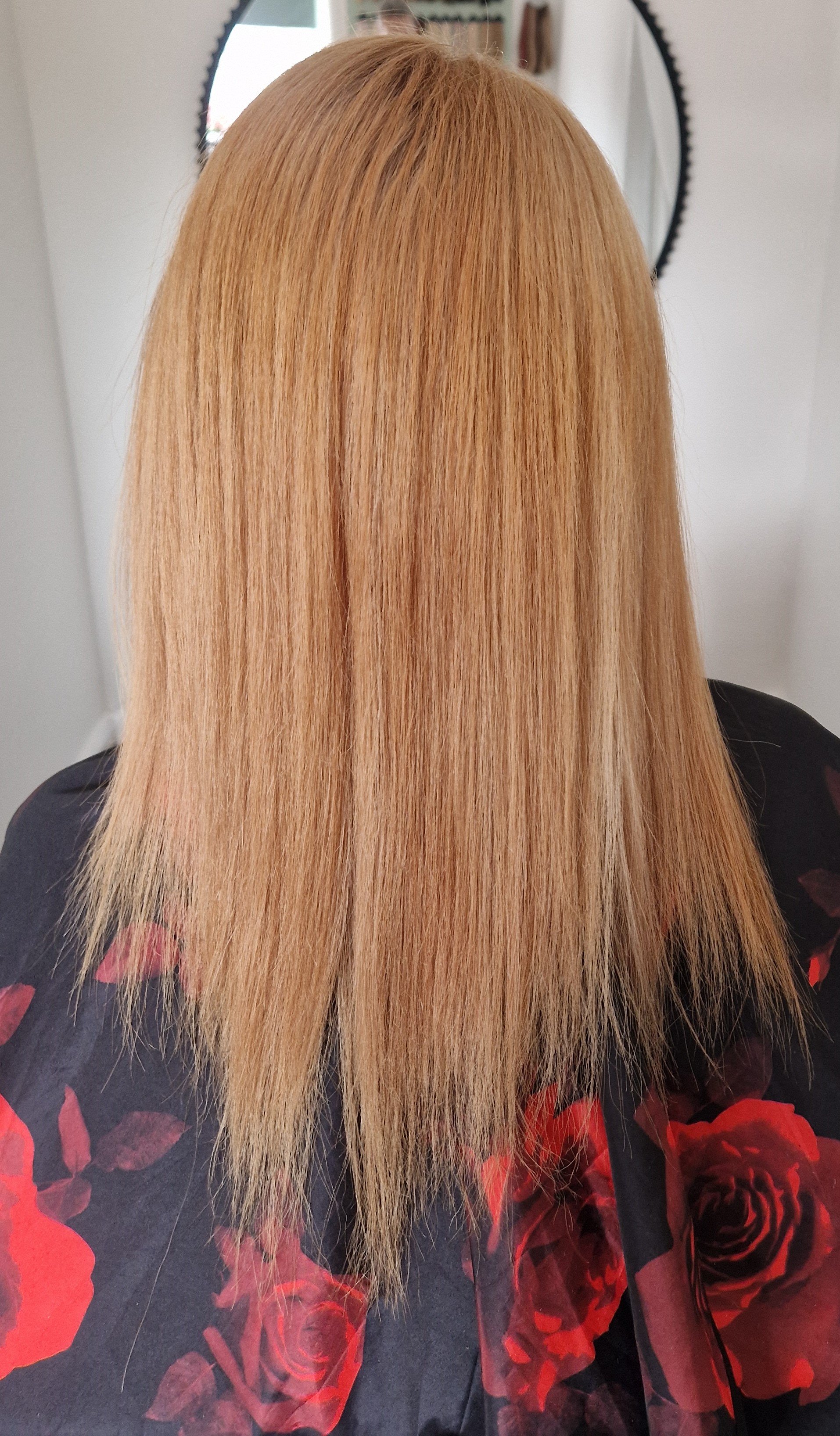 The image shows the back of a person's head with straight, long, blonde hair. They are wearing a black top with a red floral pattern.